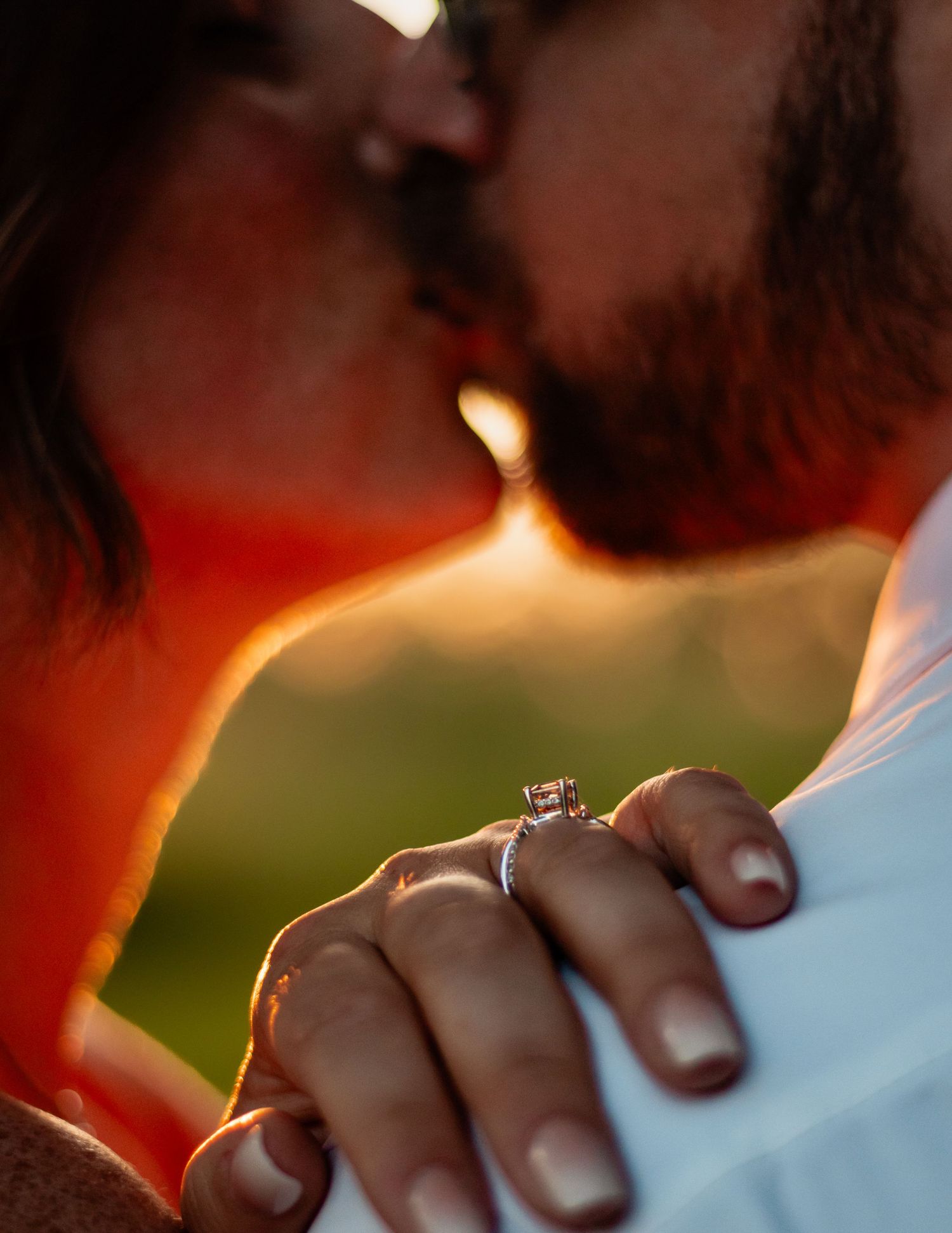 Couple kissing at golden hour with an engagement ring prominently displayed on the woman's hand in the foreground.