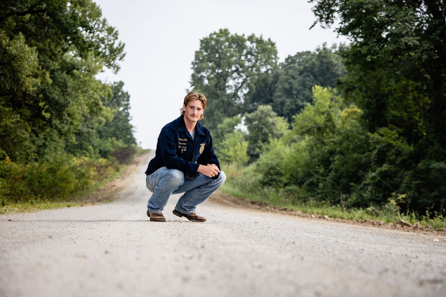 Person crouching on a rural tree-lined road wearing a dark jacket and jeans on a sunny day.
