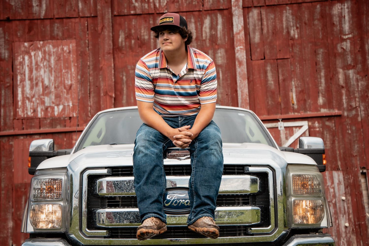 Young man in striped shirt and cap sitting on hood of Ford truck in front of red barn.