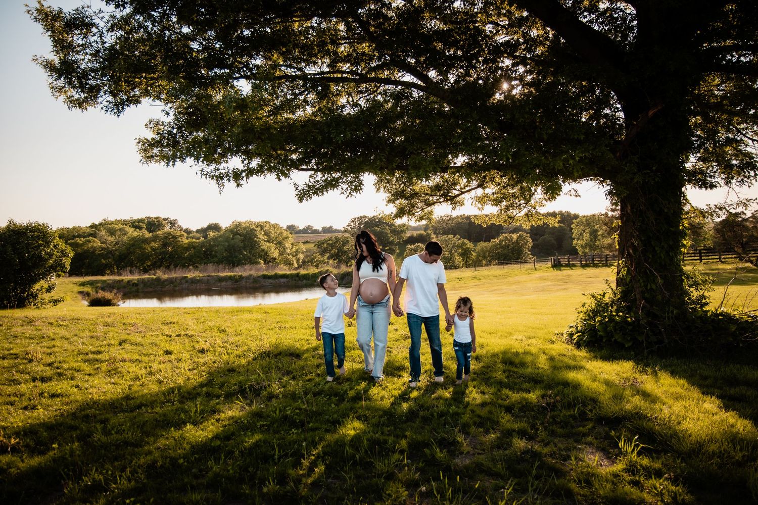 A pregnant woman holds hands with her family walking toward a pond under a large tree.