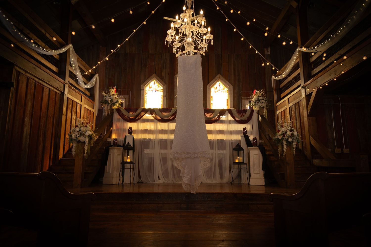 Romantic rustic barn wedding ceremony venue with chandelier, string lights, stained glass windows, and draped altar.