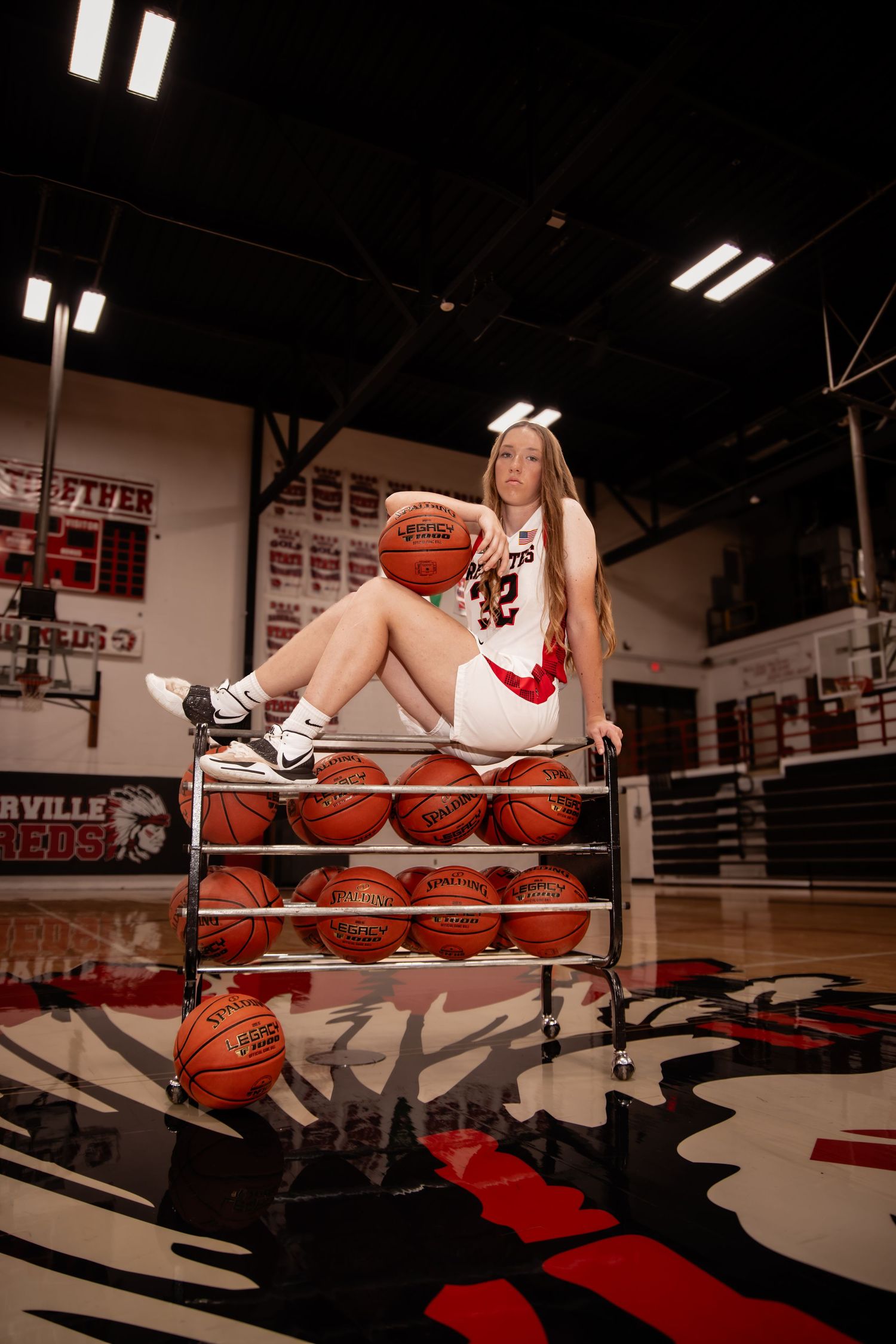 Female basketball player in white jersey #32 sits atop a ball rack in a gym, posing for senior photos.