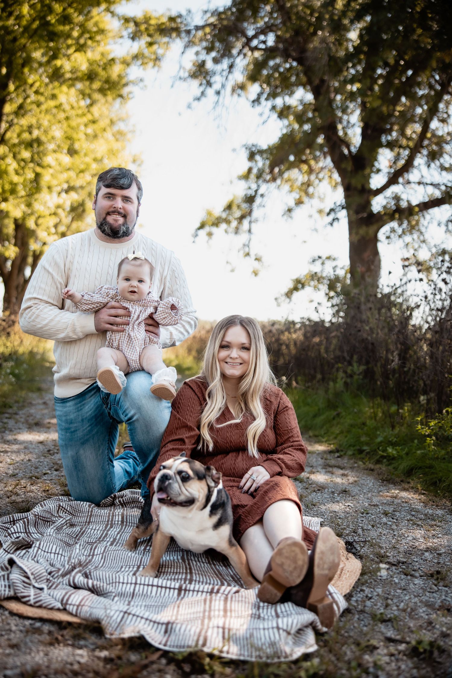 Family with baby and pug dog posing on blanket on rural dirt path surrounded by autumn trees.