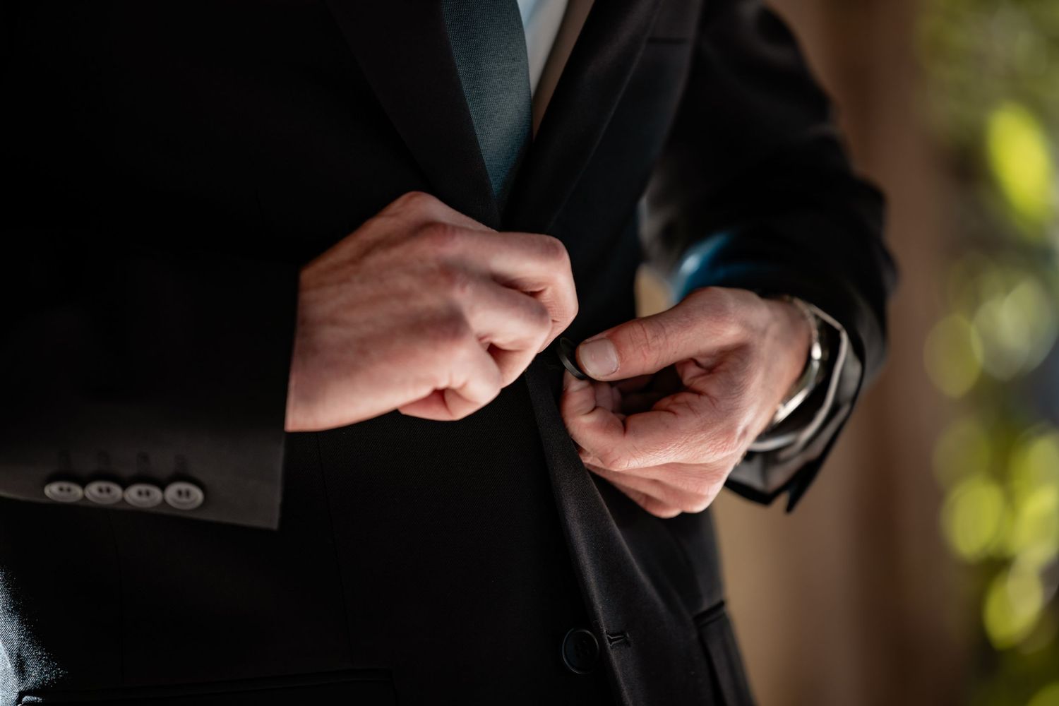 Man in black suit buttoning jacket, dressed formally for a wedding or special event.