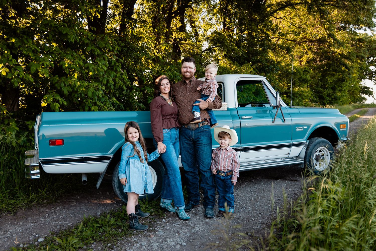 Family of five posing by a vintage teal pickup truck on a gravel path surrounded by lush green trees.