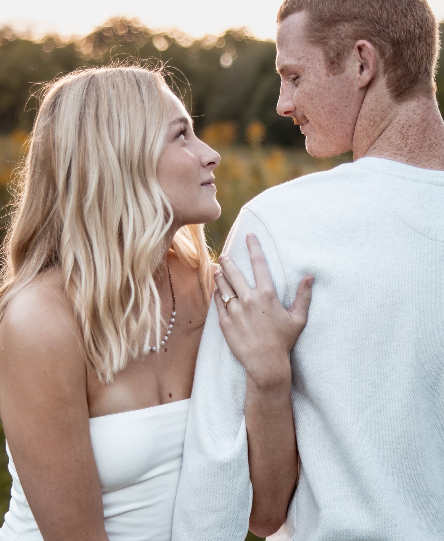 A blonde woman in white gazes at a freckled redhead man outdoors during golden hour, her hand resting on his back.