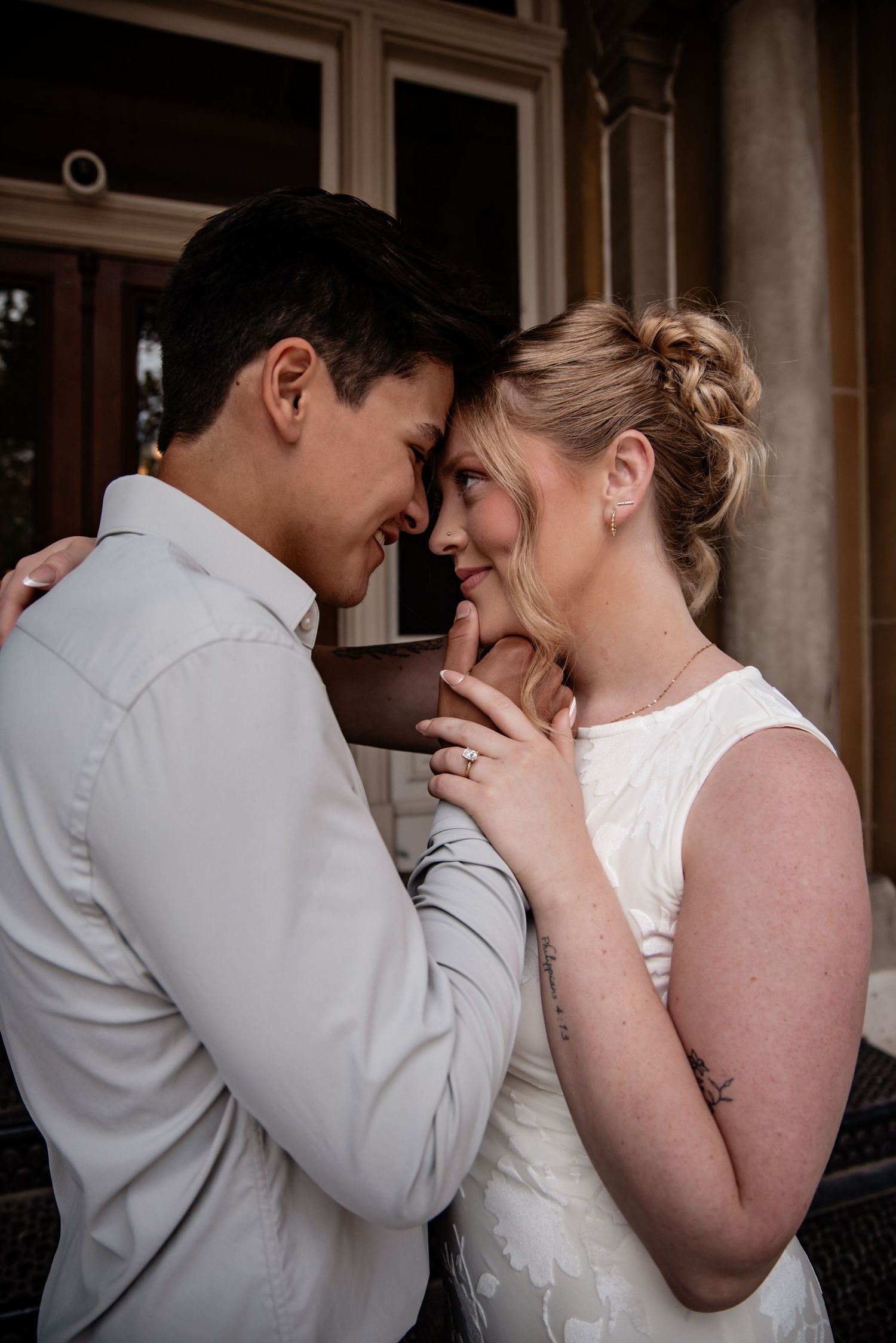 A bride and groom share an intimate forehead touch moment, smiling tenderly in wedding attire outside a building.
