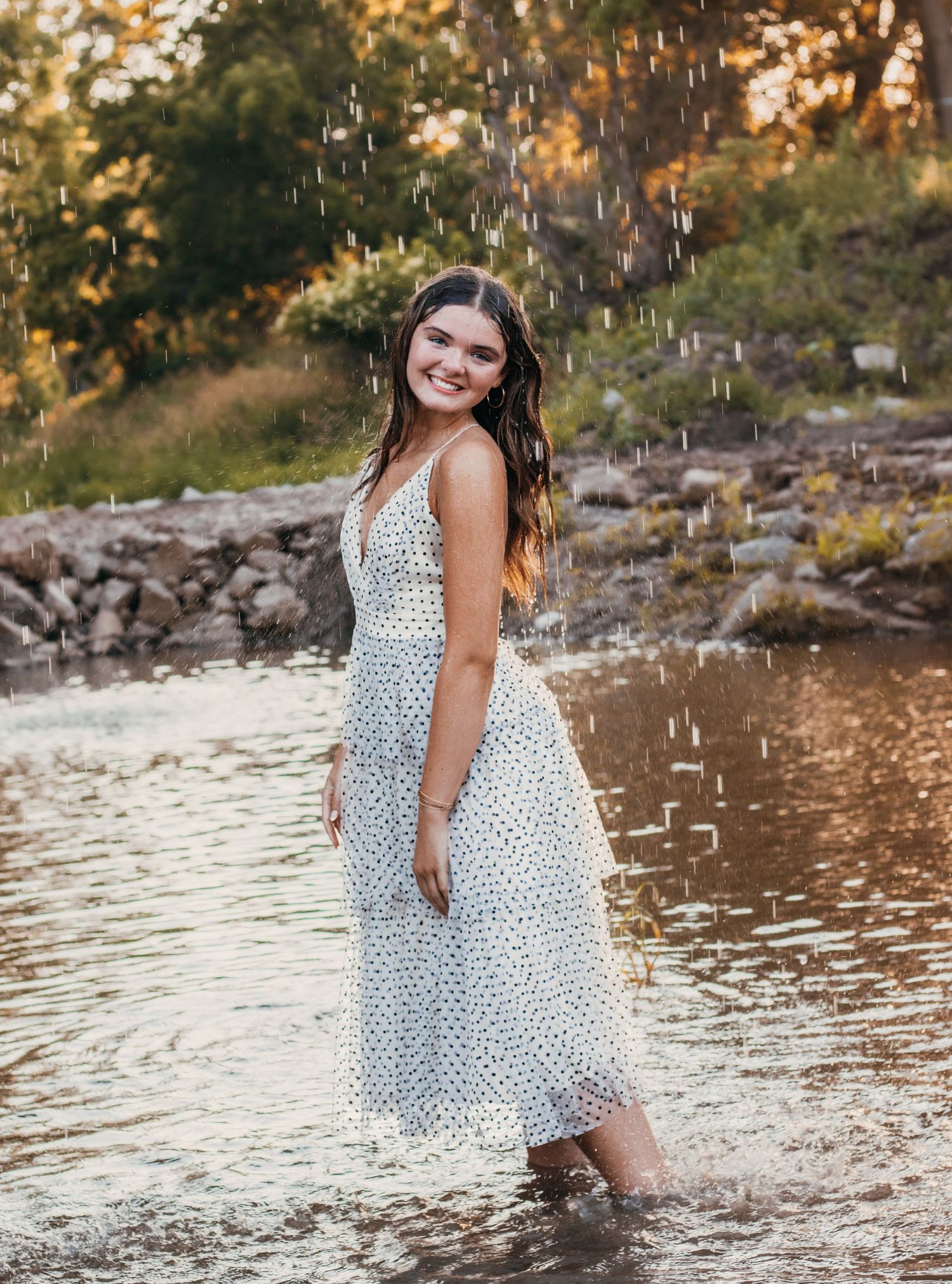 Young woman in white floral midi dress standing in shallow water during golden hour rain shower.