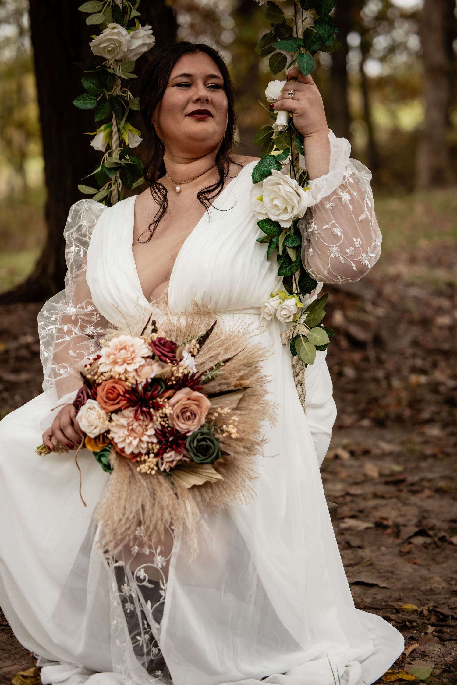 Bride in white lace gown sitting on a floral swing holding a boho bouquet in autumn woods.