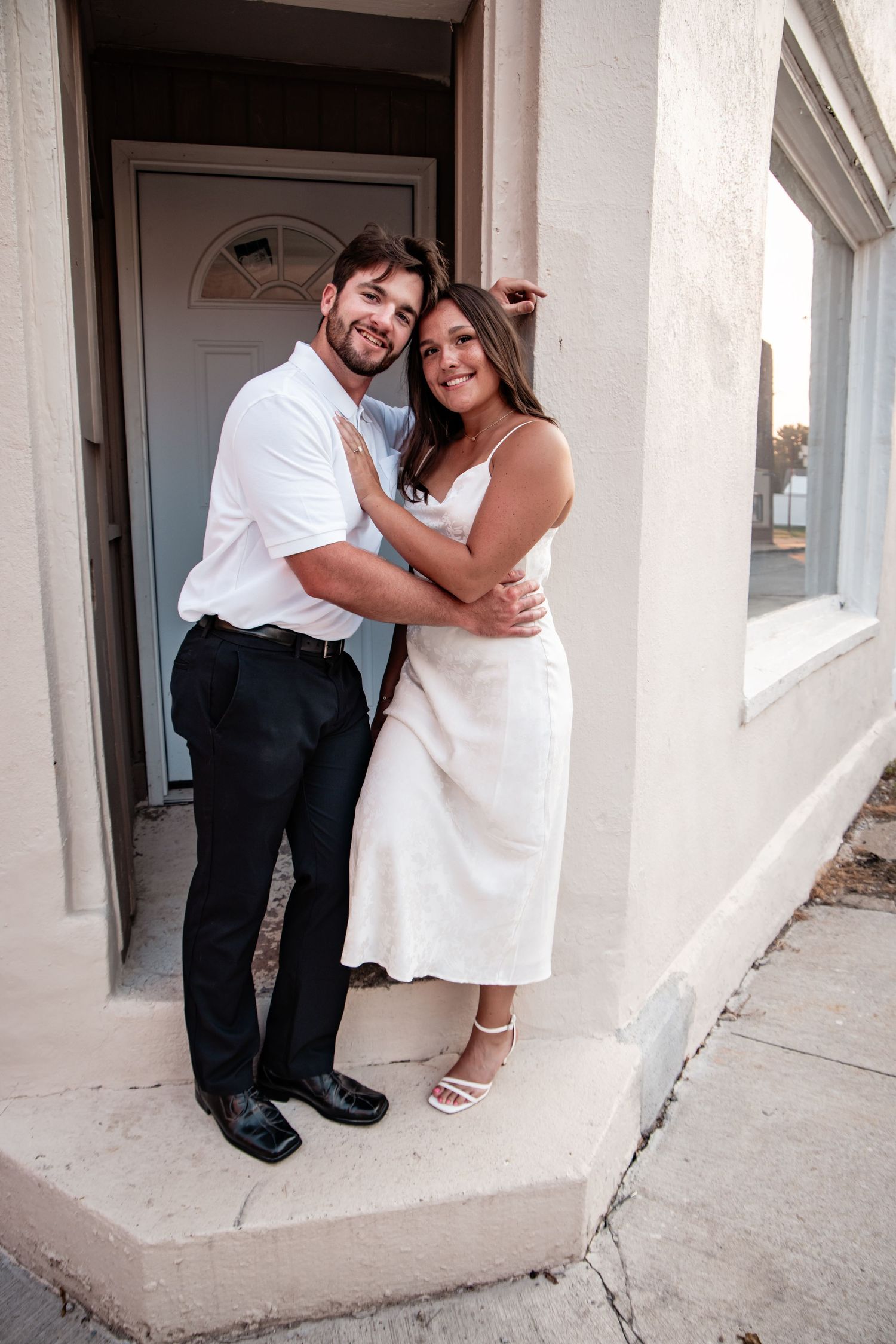 A smiling couple embraces outdoors, man in white shirt and black pants, woman in white midi dress and heels.