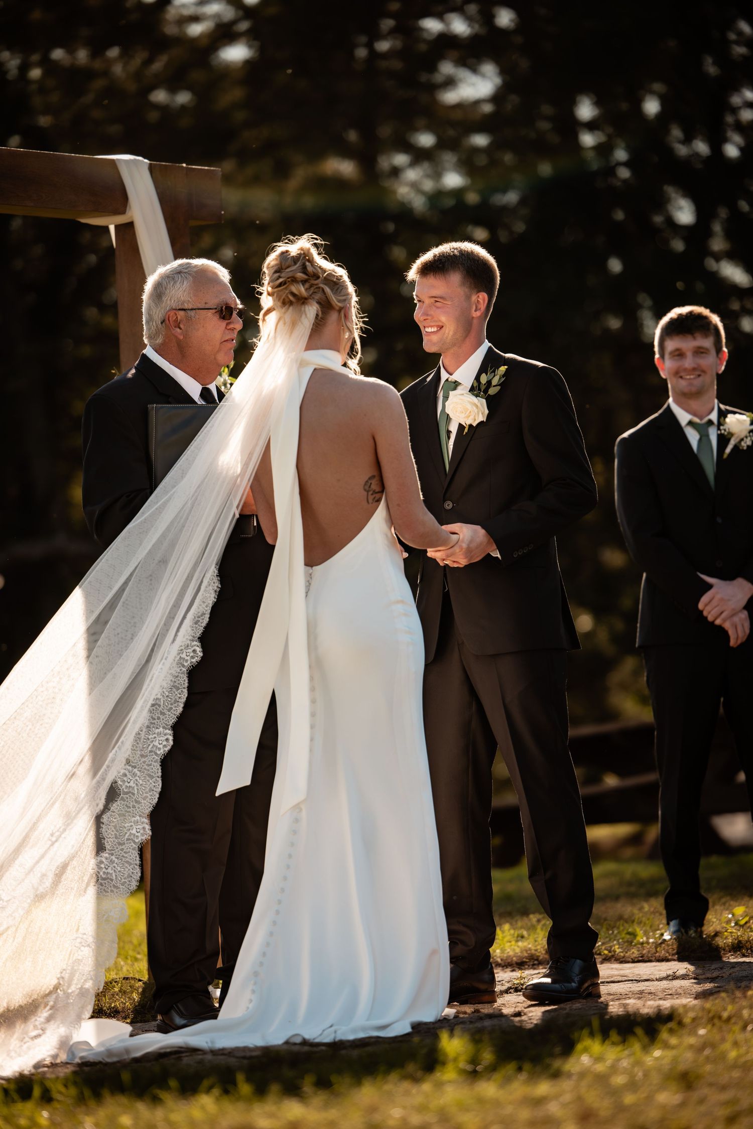 Outdoor wedding ceremony with bride in backless white gown and veil, groom in black suit smiling, officiant and groomsman present.