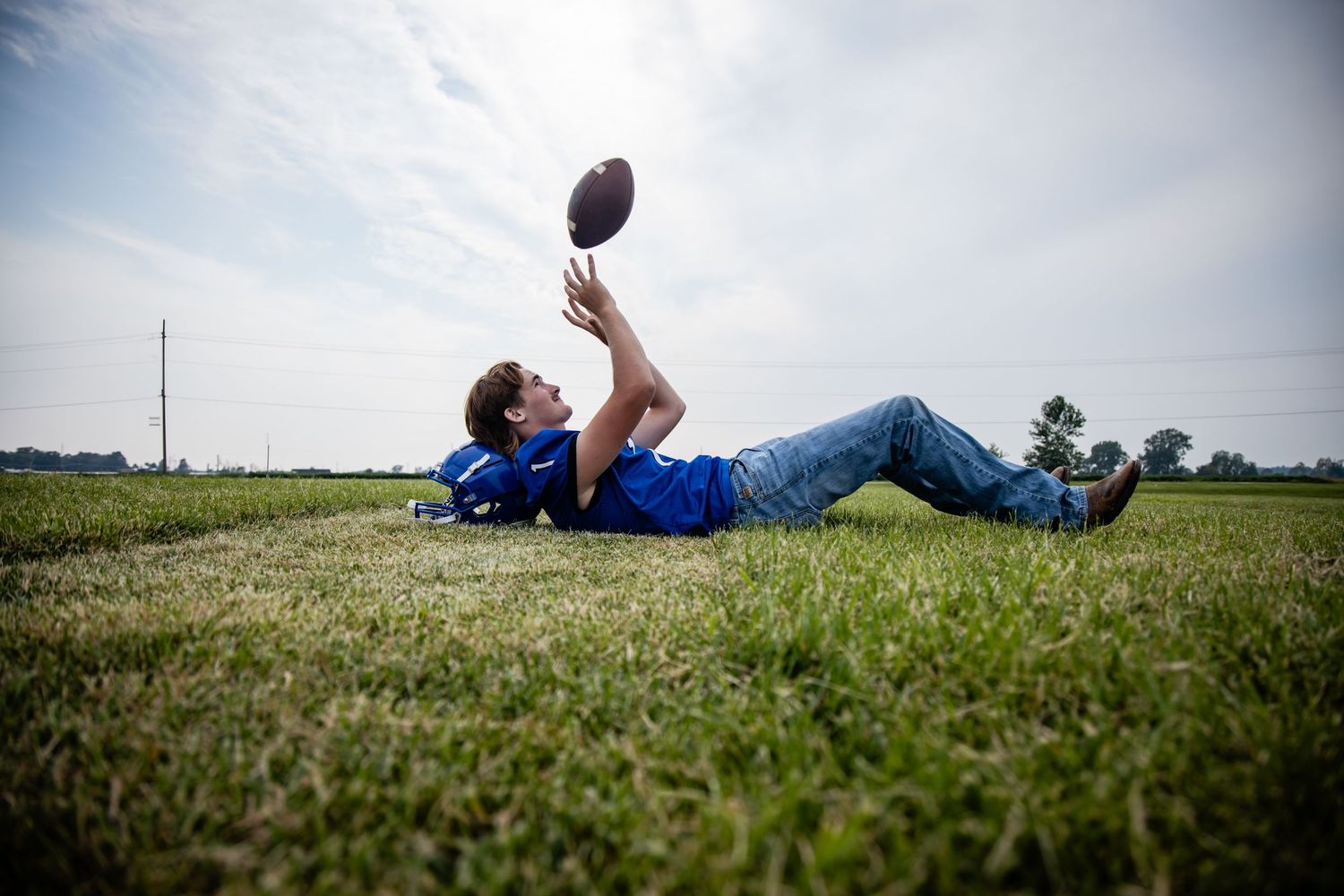 Boy lying on grass field tossing a football into the air on a cloudy day.