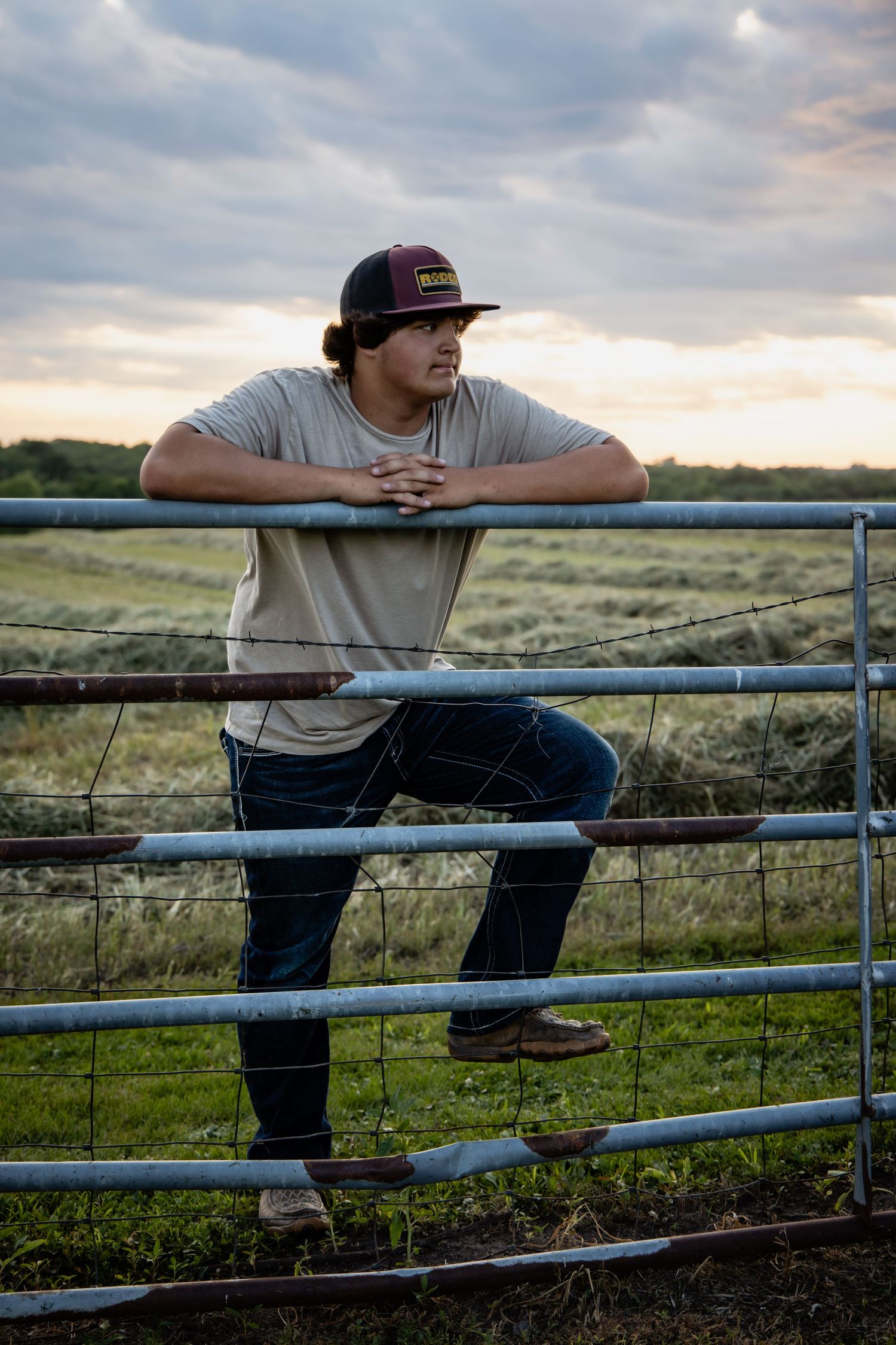 Young man in cap and jeans leans on a metal farm gate at sunset with green fields in the background.