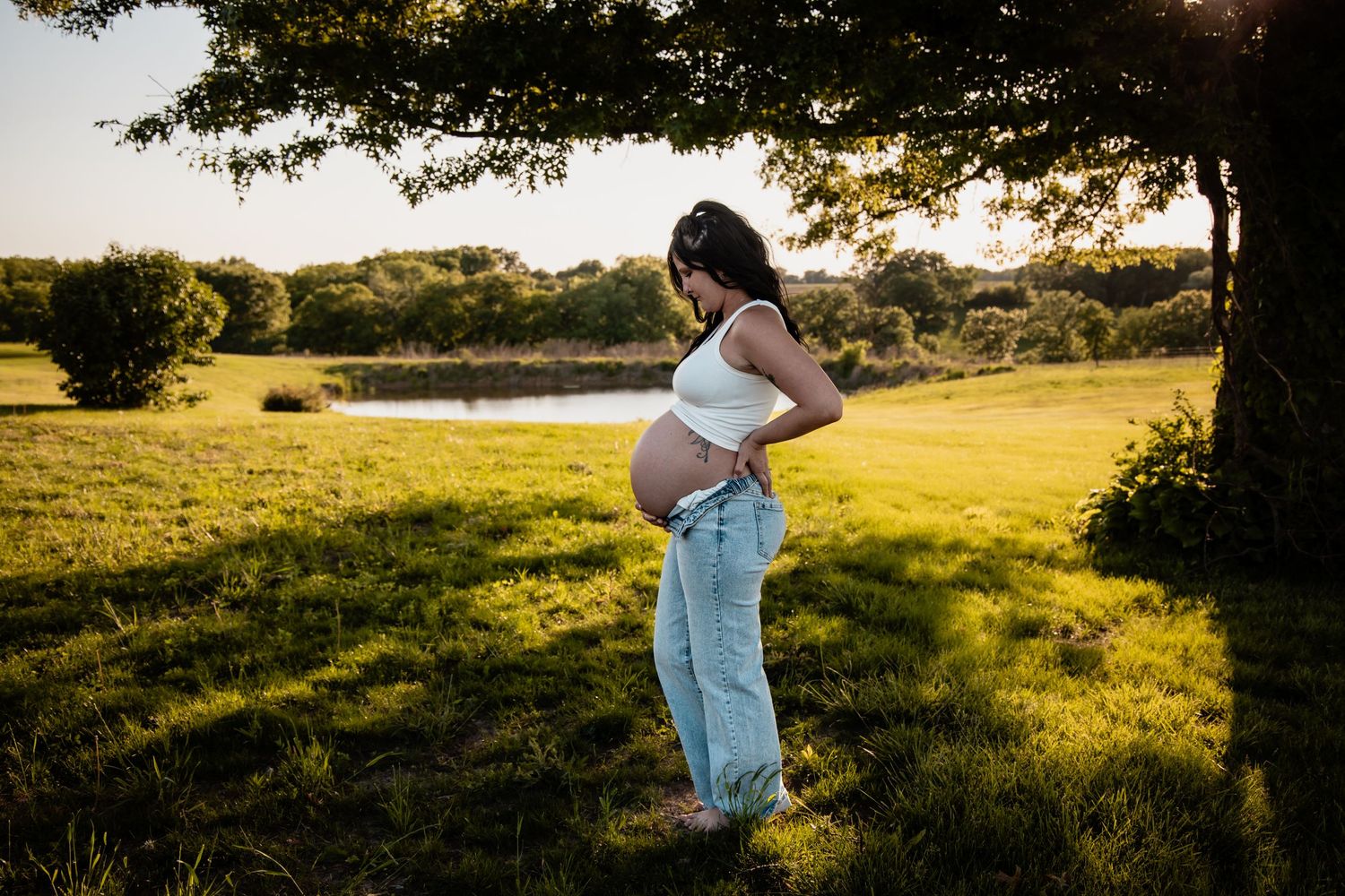 A pregnant woman in a white crop top and jeans stands barefoot in a sunny green field.