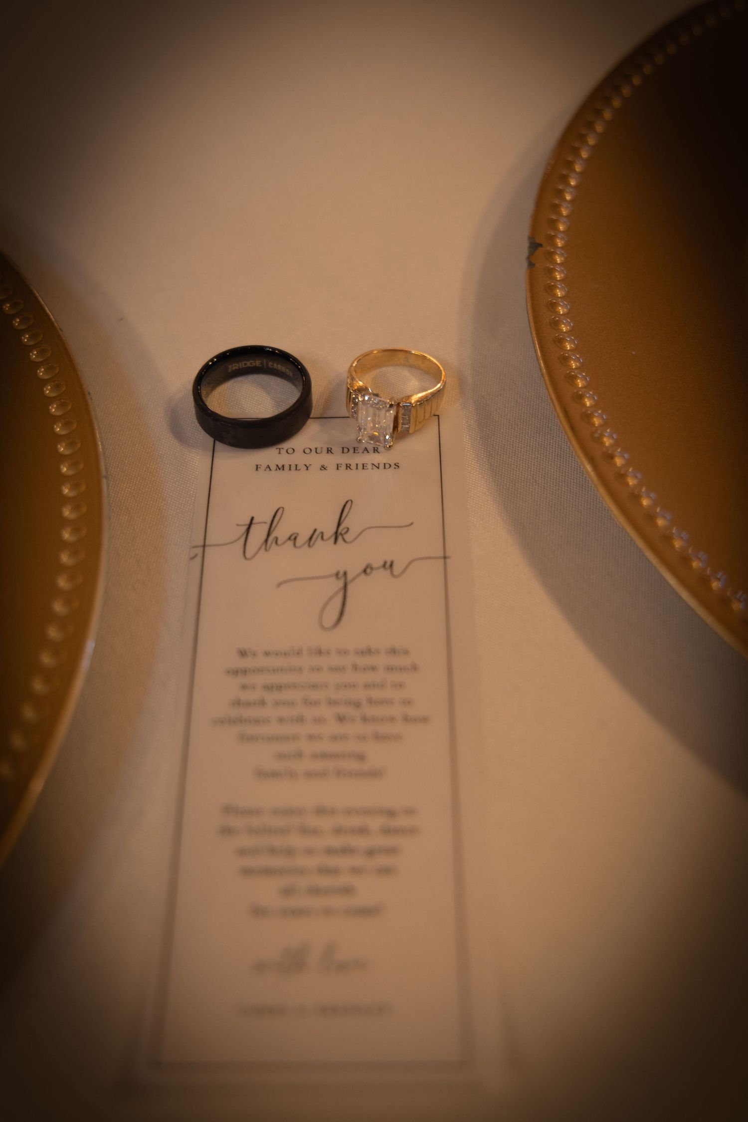 Wedding rings, black band and gold diamond ring, resting on a thank you card between gold charger plates.