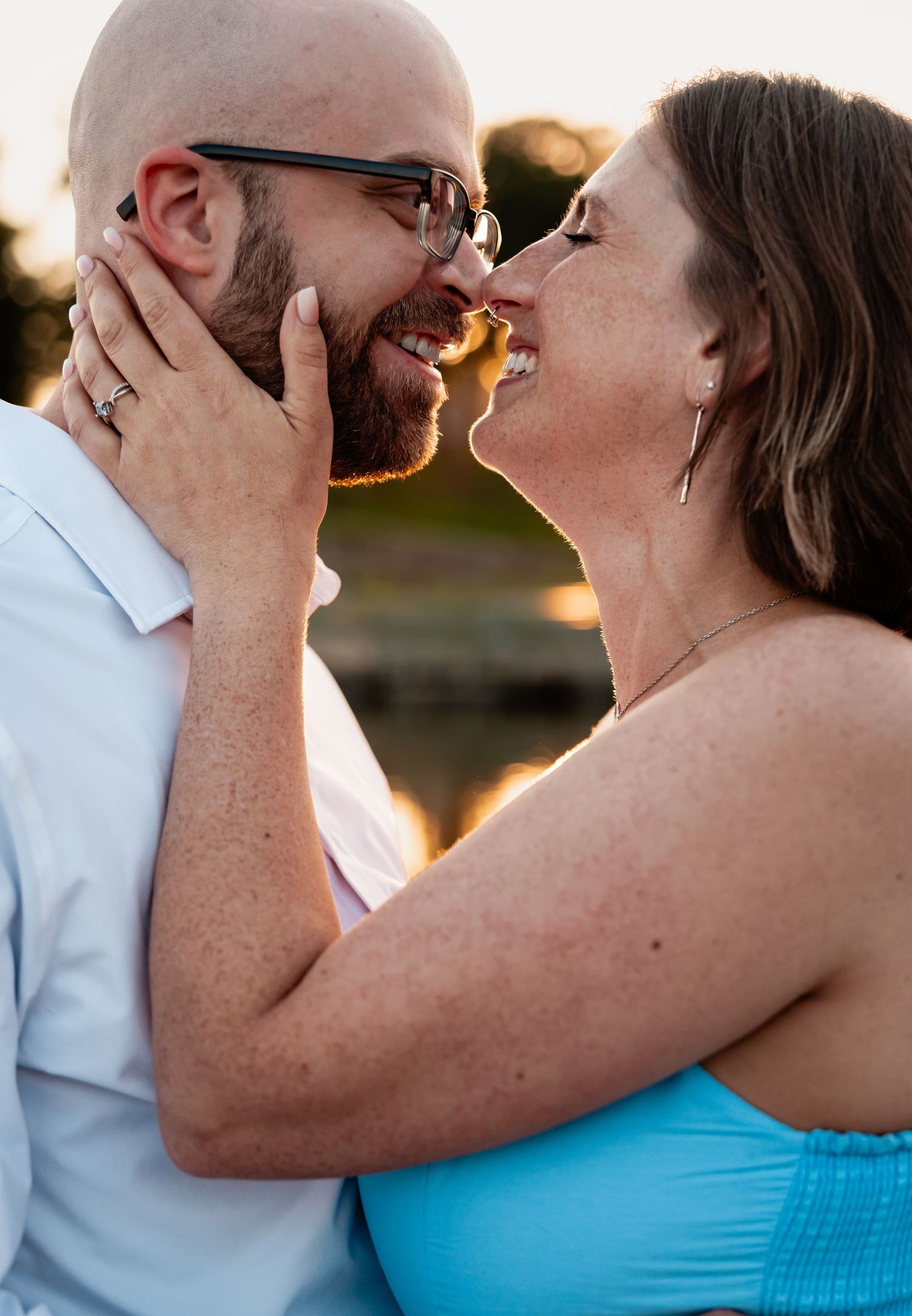 A couple shares a romantic moment, nearly kissing at golden hour, woman in turquoise top holding mans face, engagement ring visible.