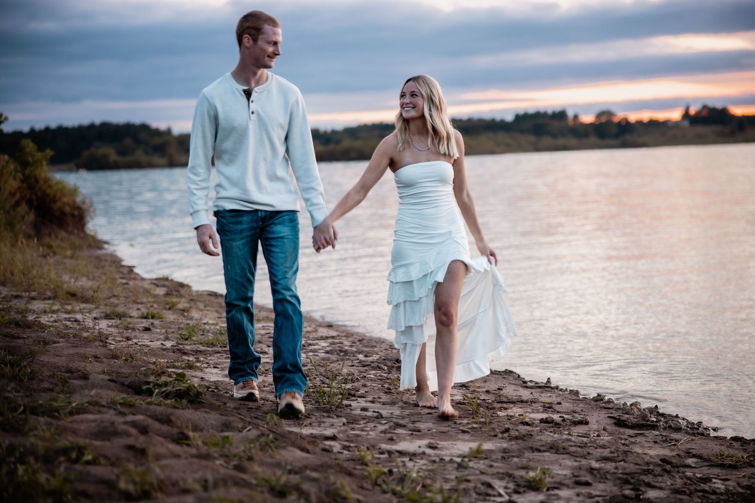 Couple in white outfits walking hand-in-hand along a sandy riverbank at sunset.