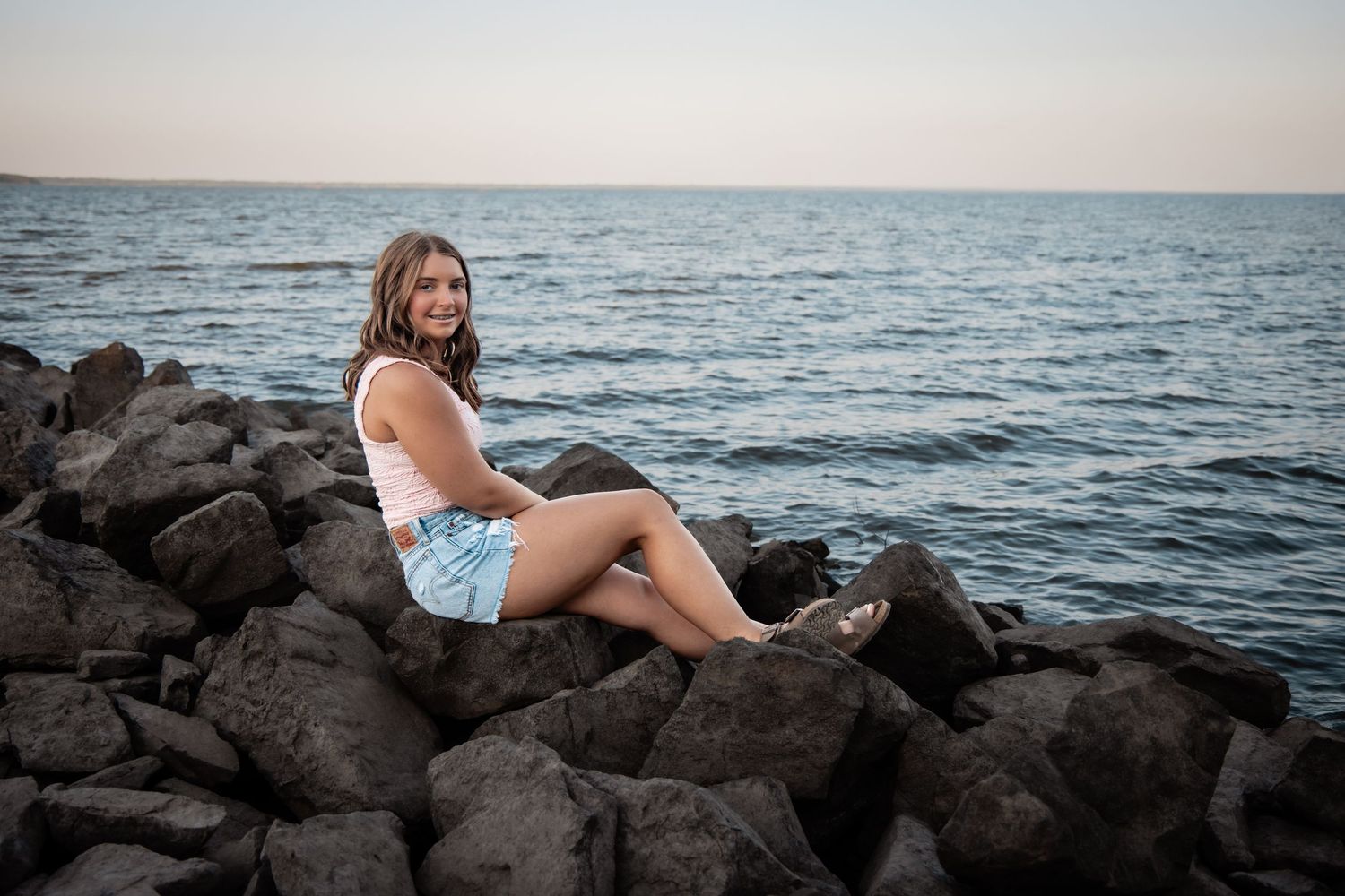 Young woman in white top and denim shorts sitting on rocky shoreline with calm ocean at sunset.