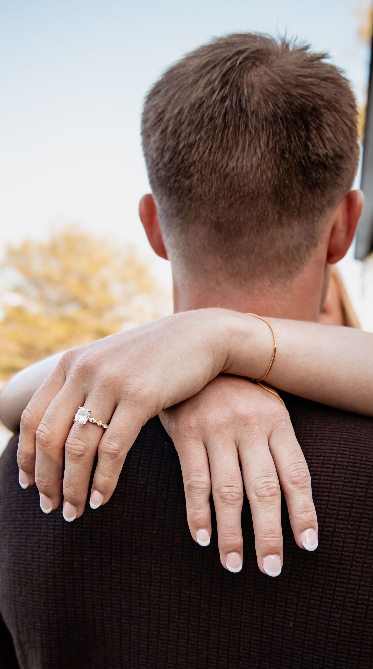 Close-up of a woman's hand with a diamond engagement ring resting on a man's shoulder from behind.