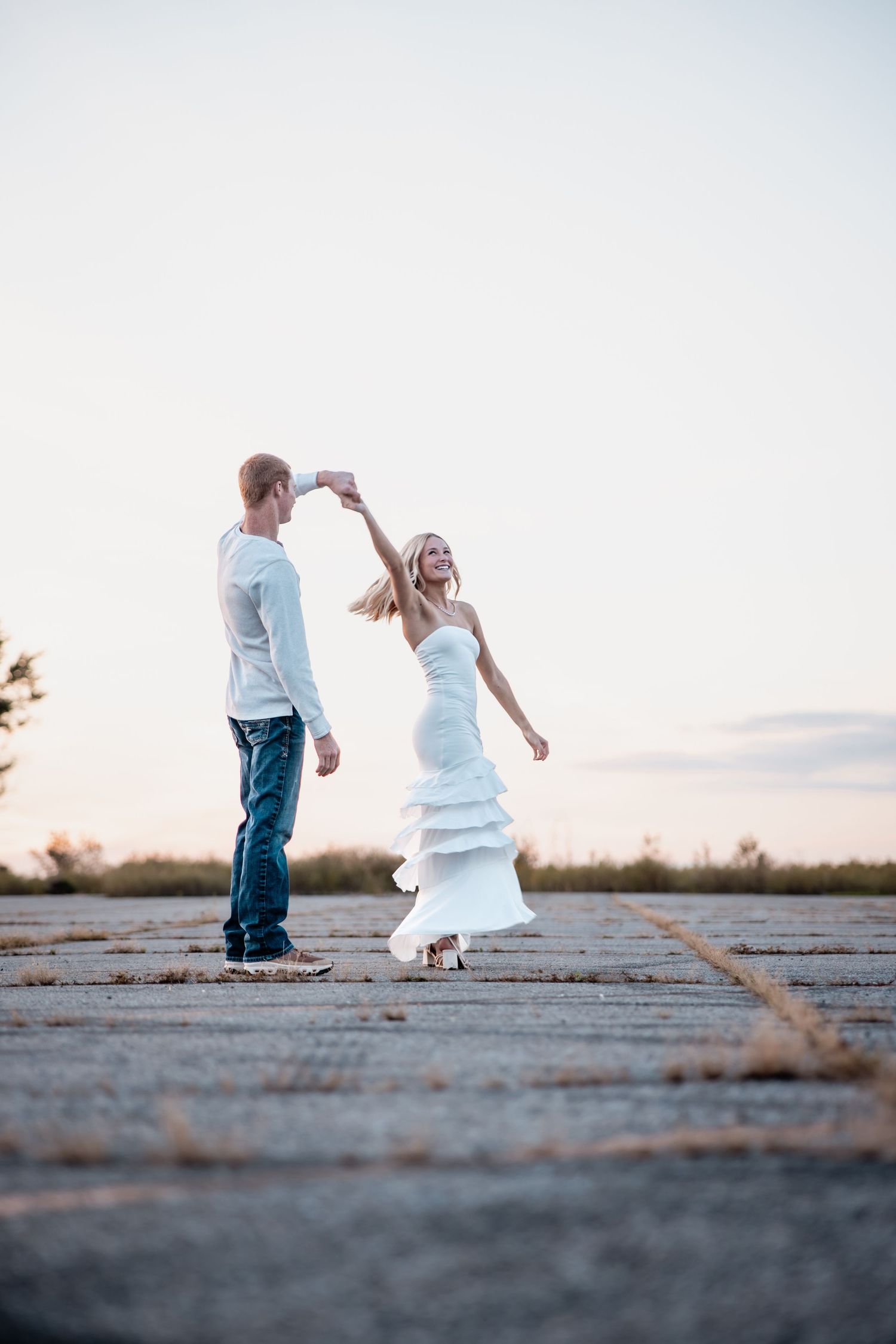 Bride in white dress dancing with groom in jeans at sunset on open pavement during outdoor wedding.