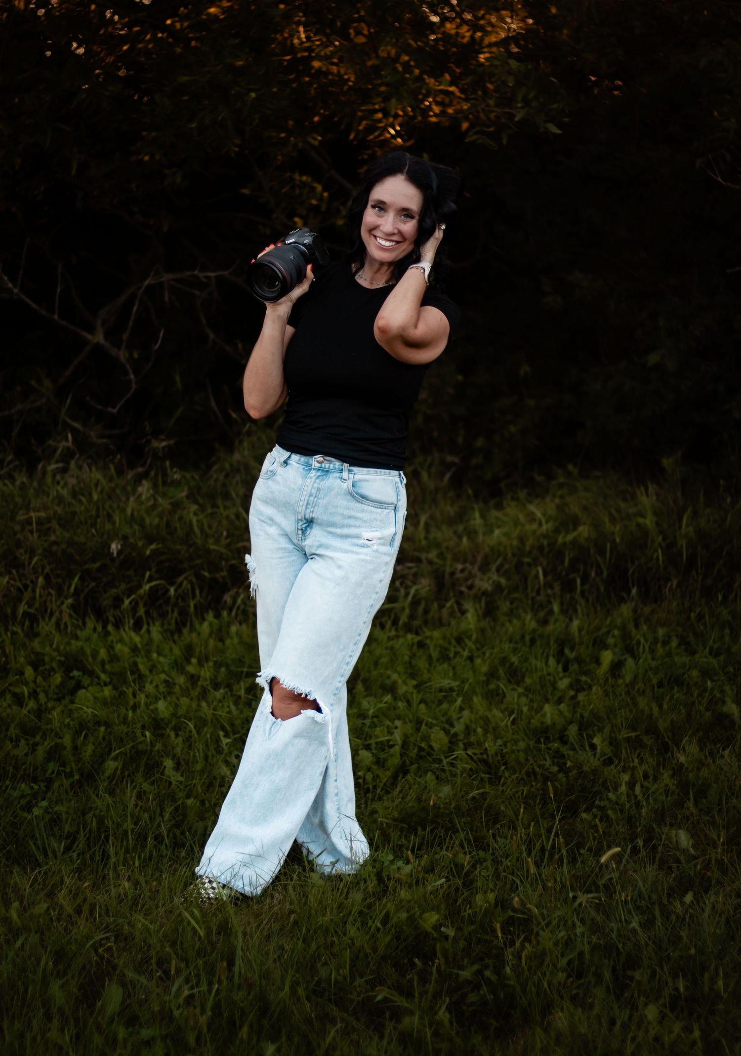 Smiling woman in black top and ripped flare jeans holds a camera outdoors in a lush green field at dusk.