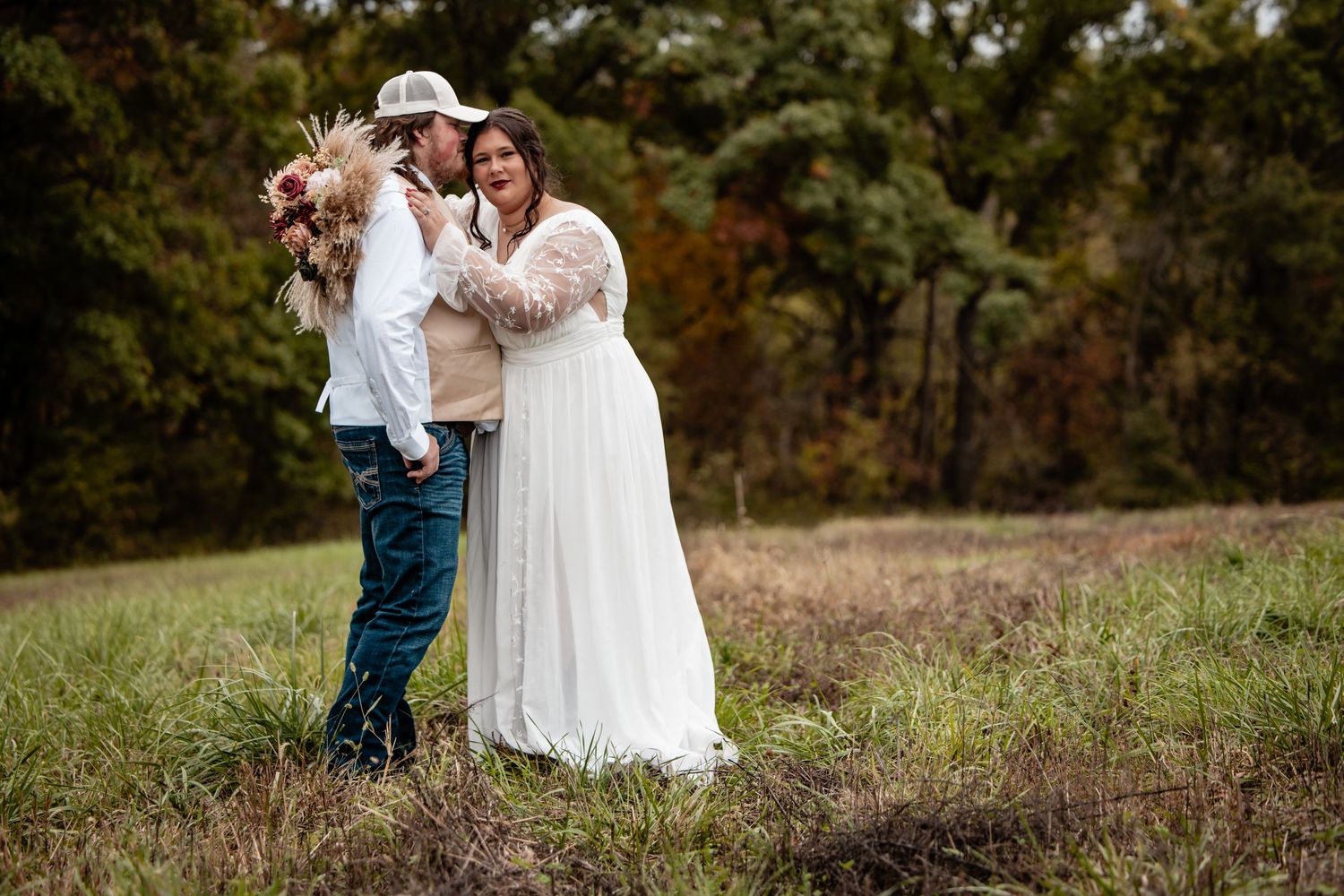 Couple in wedding attire embracing outdoors in a field with autumn trees, bride in white lace gown and groom in casual hat.