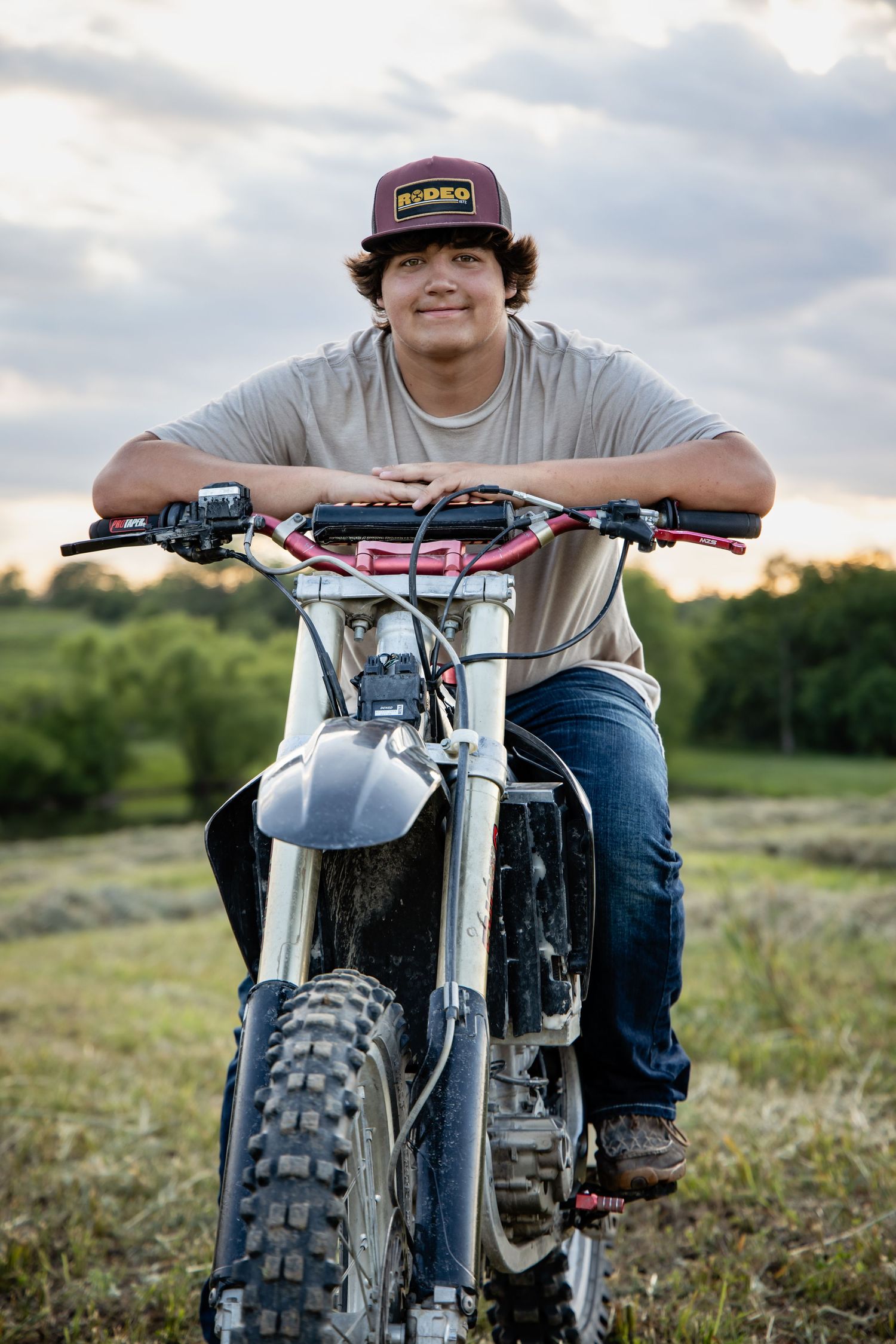 Young man in a cap rests on a dirt bike in a green field at sunset, smiling confidently.