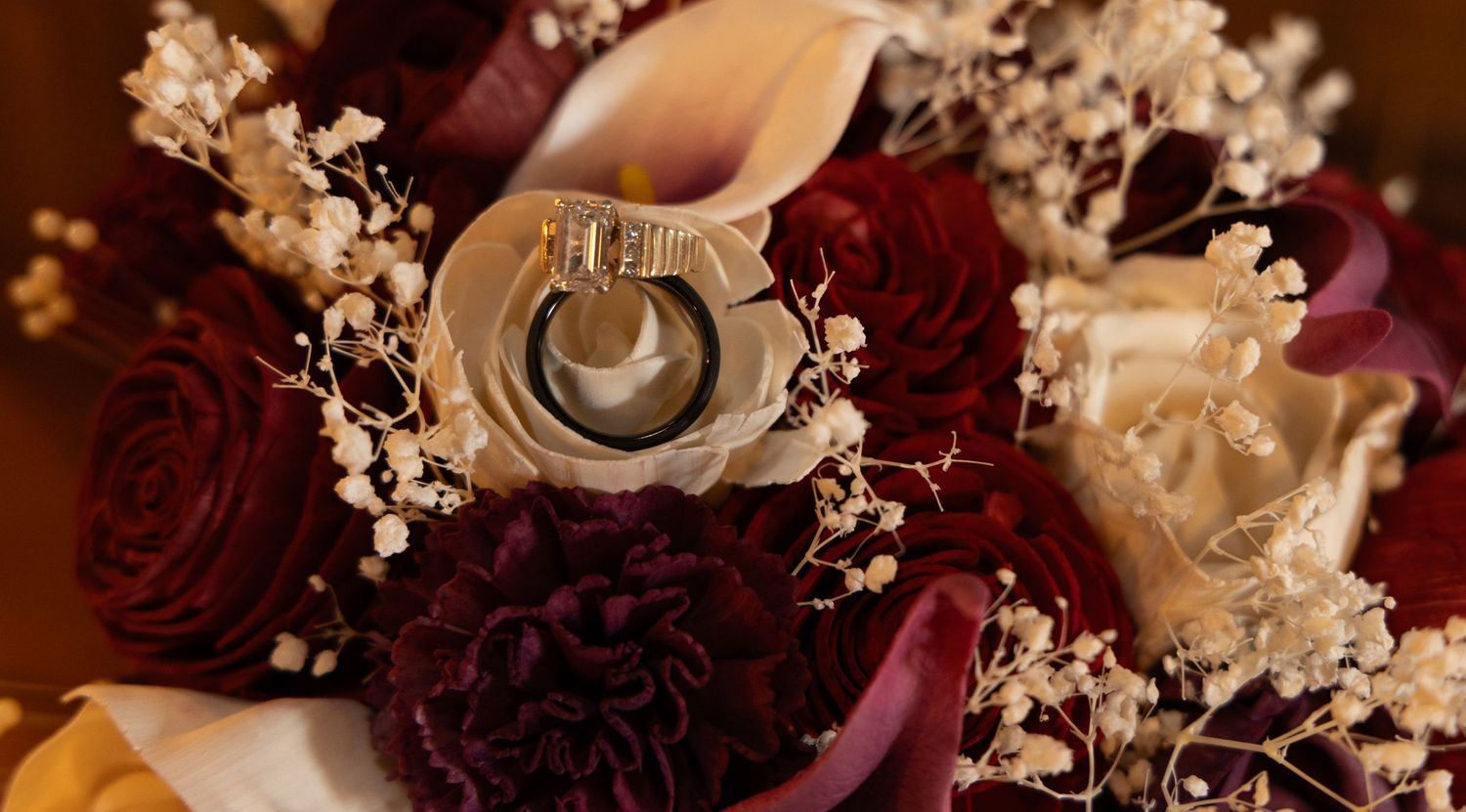 Close-up of wedding rings nestled in a bridal bouquet with deep burgundy roses, calla lilies, and baby's breath.