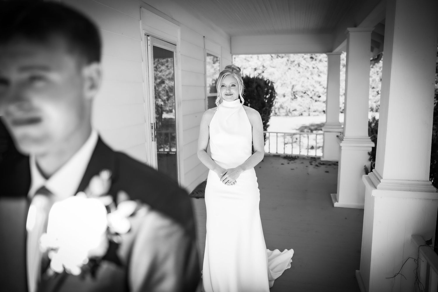 Black and white photo of a bride in a white gown smiling at groom on a porch.
