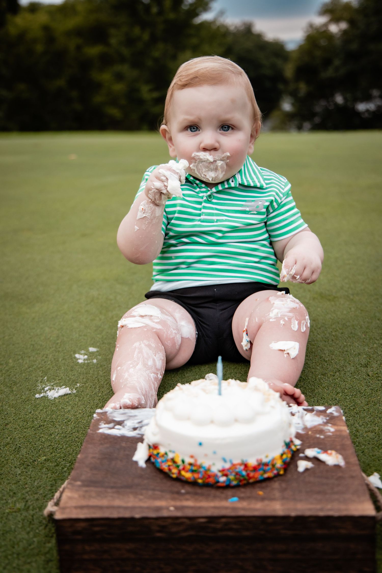 Baby boy in green shirt smashes first birthday cake outdoors on grass, face and hands covered in white frosting.