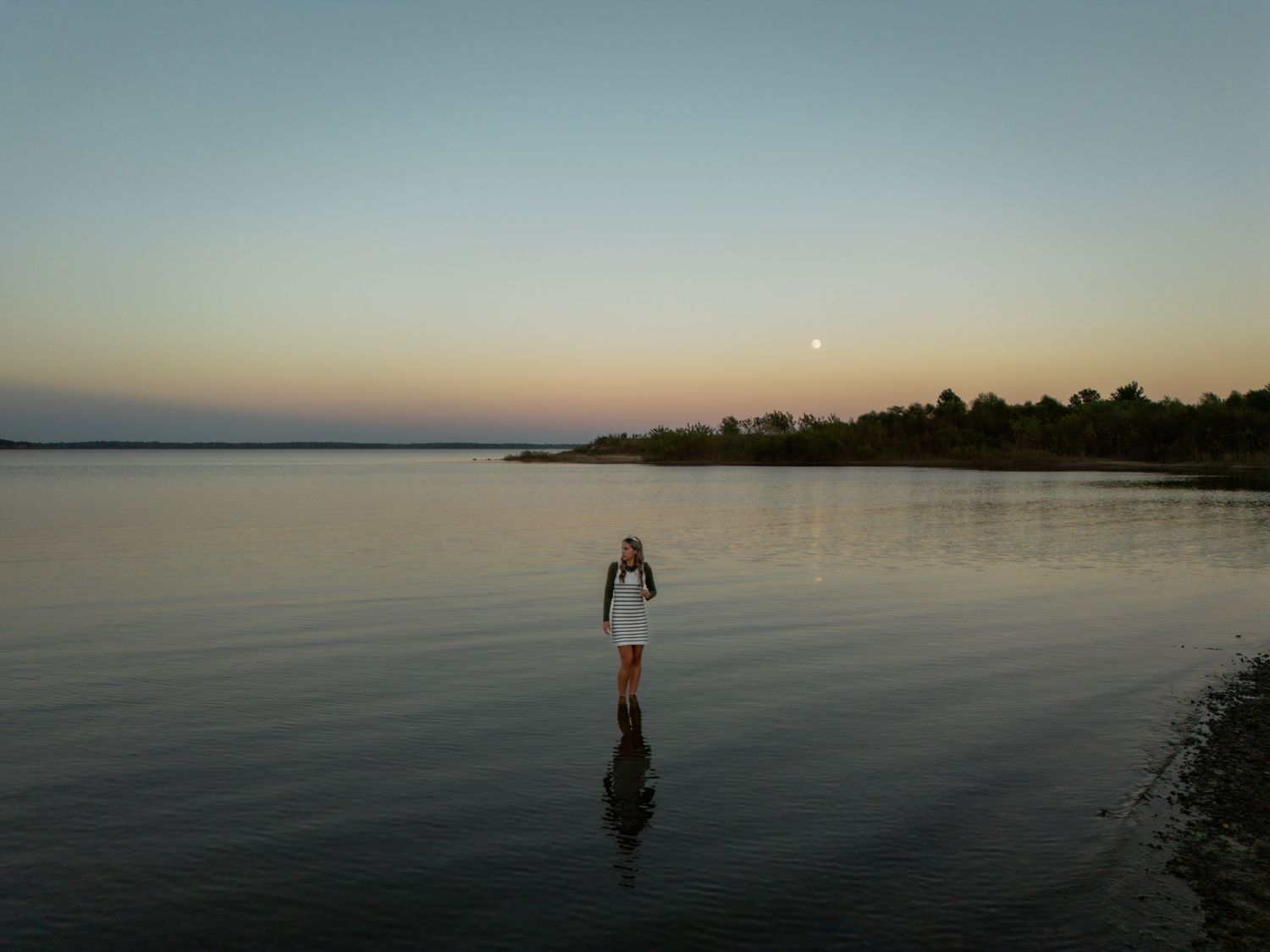 Person standing in calm shallow water at sunset with moon visible in pastel sky and tree line reflecting on still water.