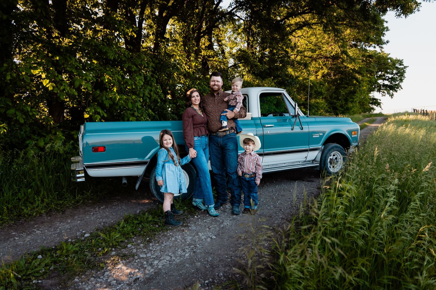 Family of five posing on a dirt road beside a classic teal pickup truck.