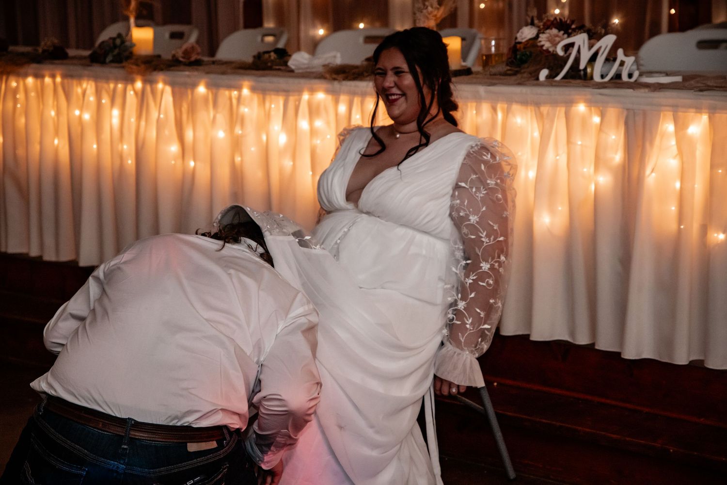 Bride laughing joyfully as groom removes her garter during lively wedding reception celebration.