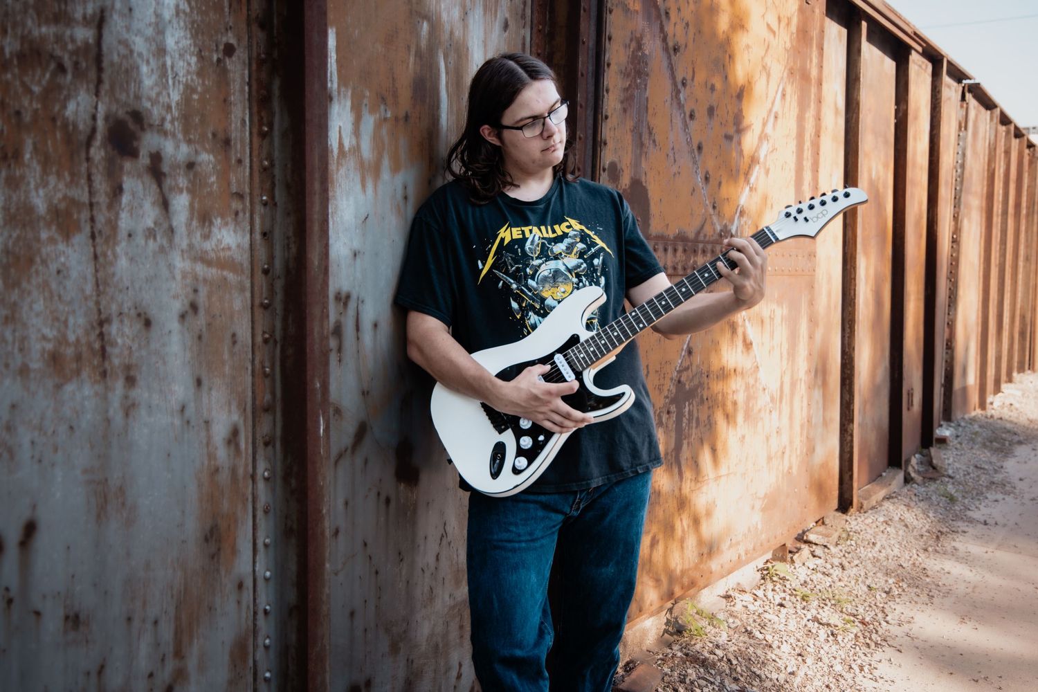 Young guitarist in Metallica shirt playing white electric guitar outdoors against rusty metal wall.