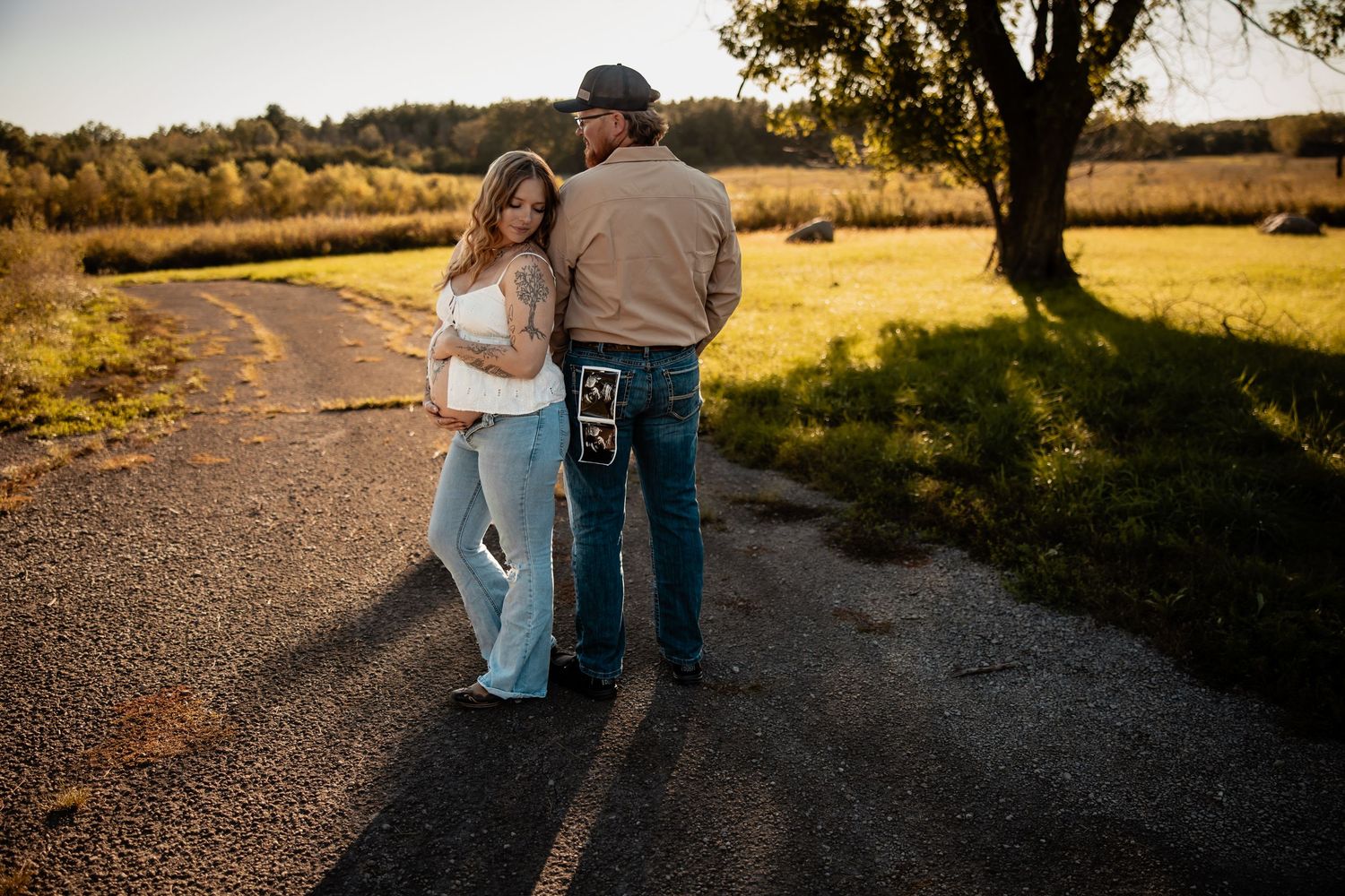 Couple standing on a rural road at golden hour, woman in white top and jeans leaning on man in tan shirt and jeans.