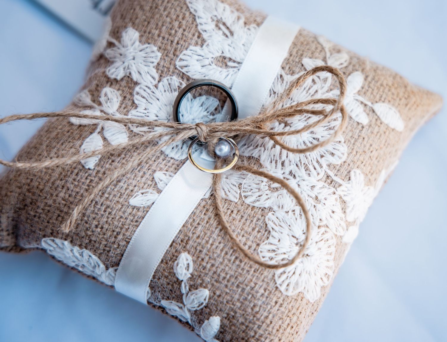 Rustic burlap ring bearer pillow with white lace, ribbon, and twine bow holding two wedding rings.