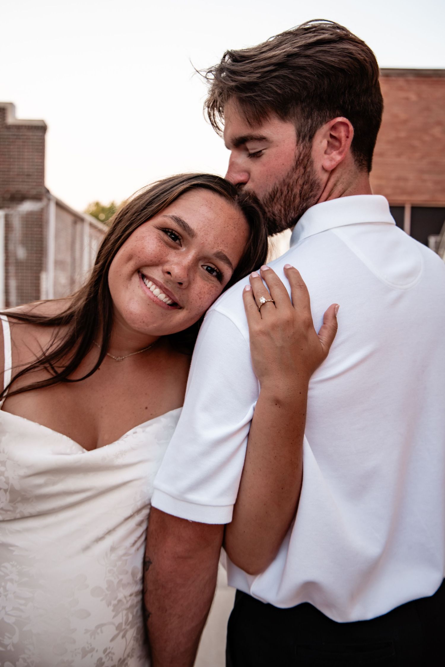 Bride in white dress smiles as groom kisses her cheek, showing engagement ring outdoors.