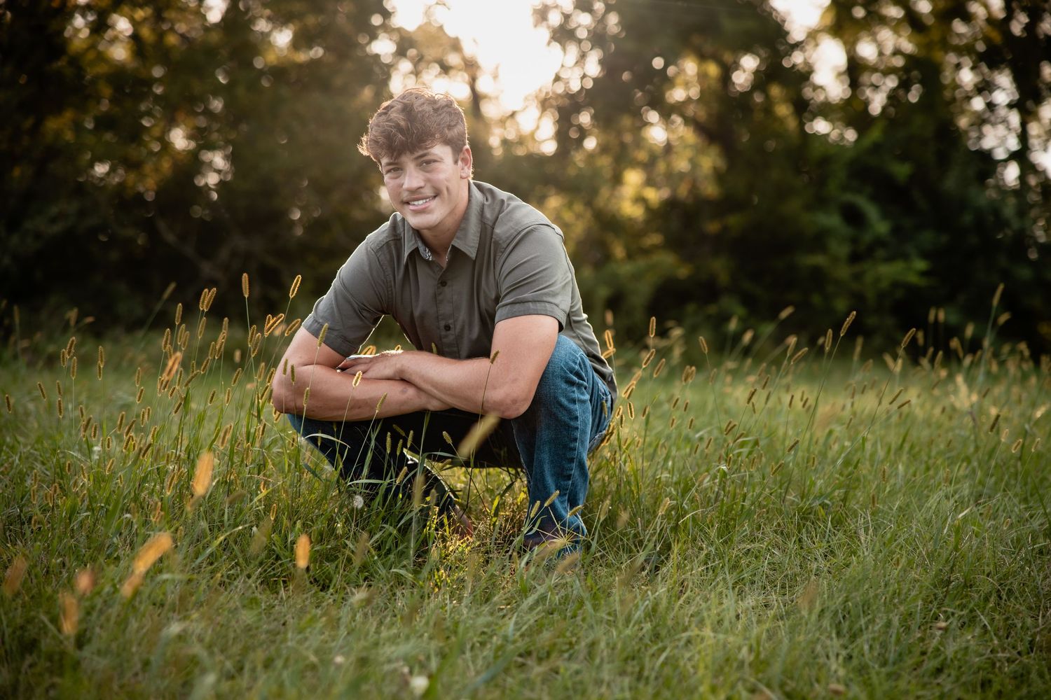 Young man crouching in a sunlit grassy field, smiling, wearing a gray polo and jeans, with trees in the background.