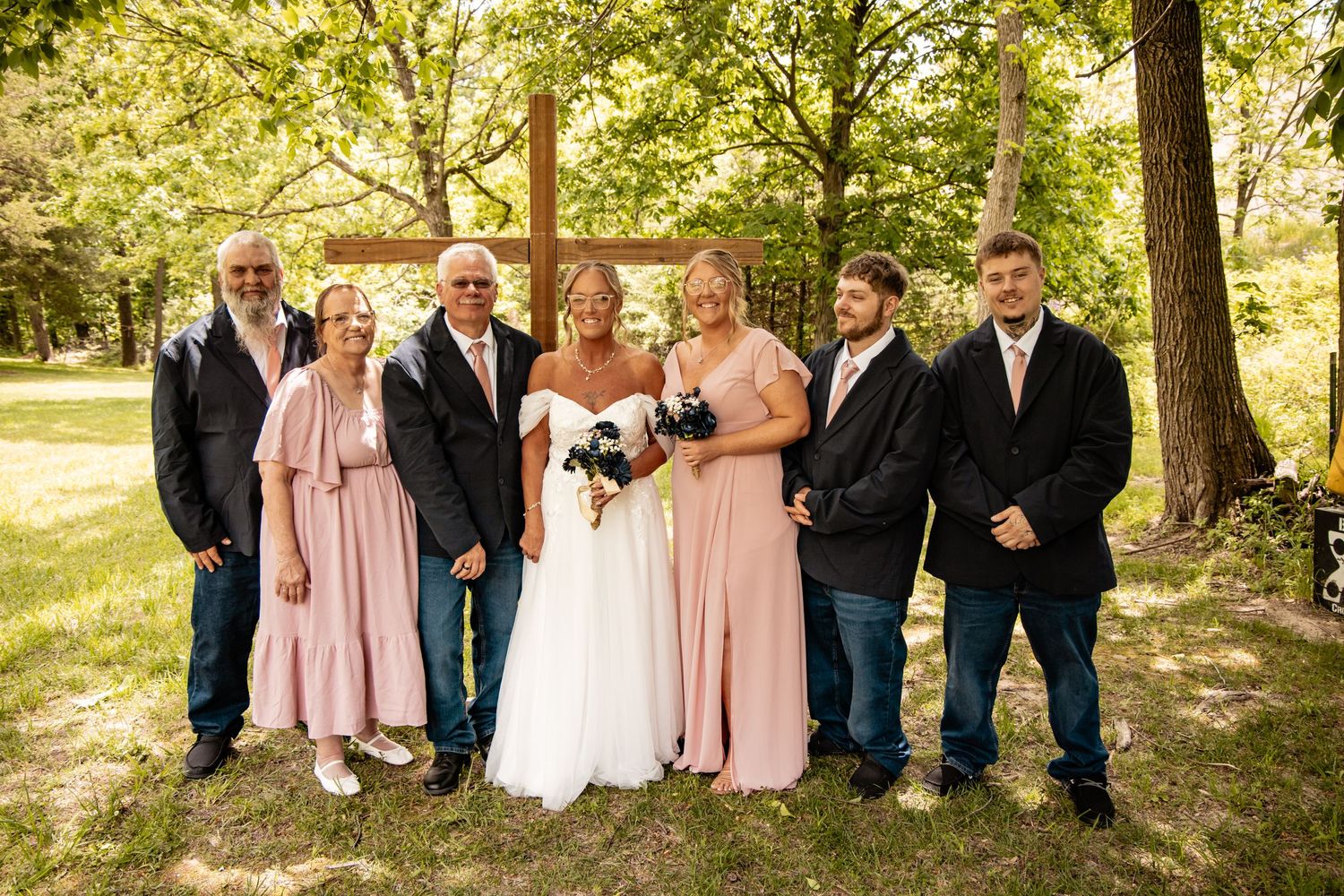 Wedding party poses together in front of a wooden cross, bridesmaids wearing blush pink dresses.