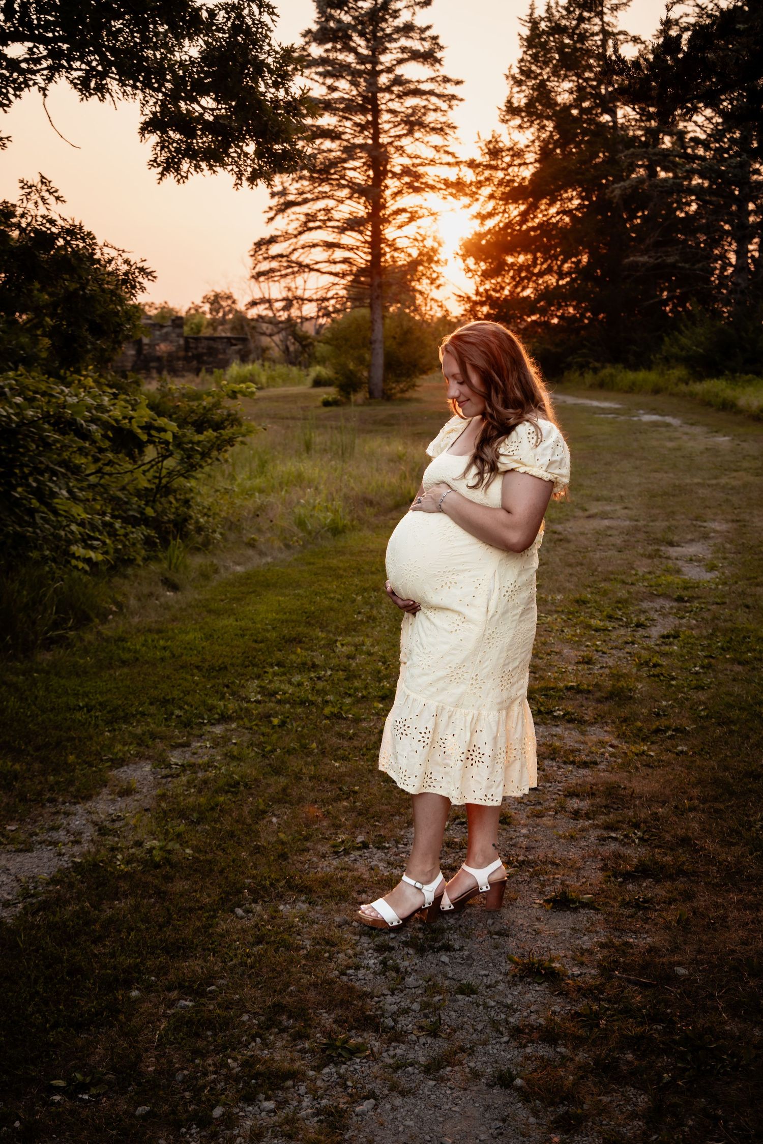 Pregnant woman in white lace dress holding belly at golden hour sunset in lush green park setting.