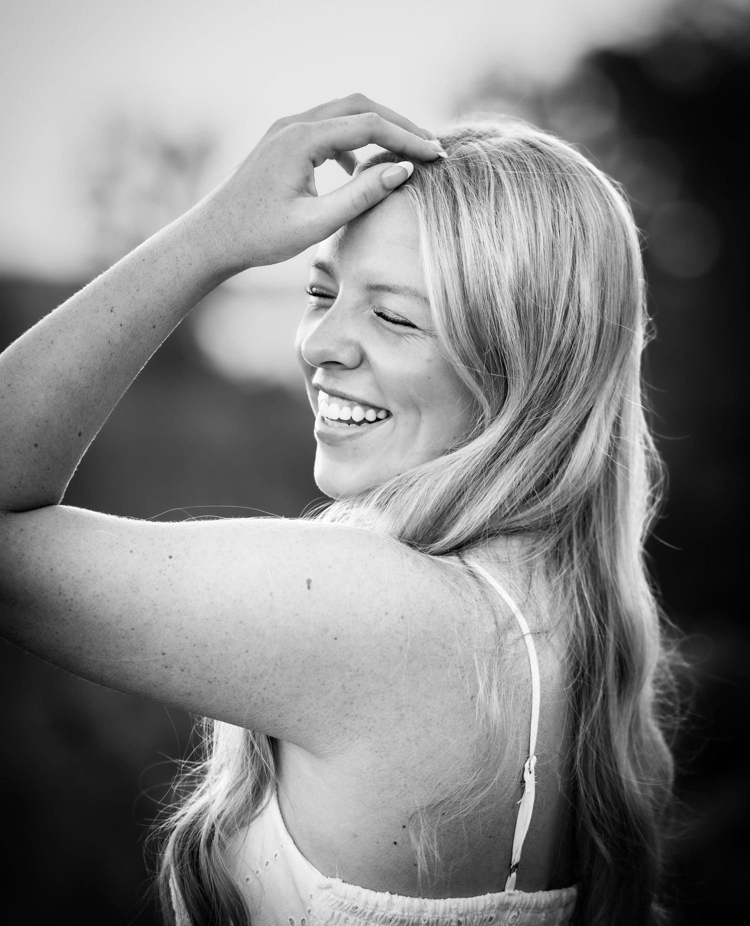 Black and white portrait of a laughing blonde woman with hand on forehead, enjoying a carefree outdoor moment.