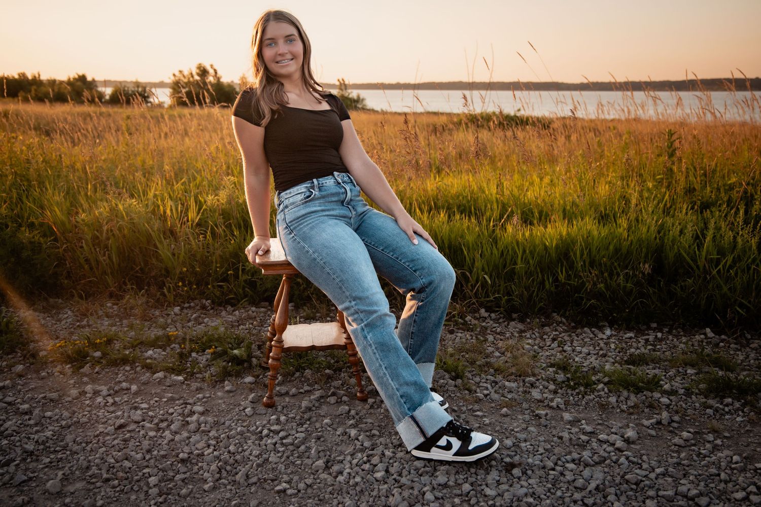 Young woman in jeans and black top sitting on a wooden chair outdoors at golden hour near a lake.