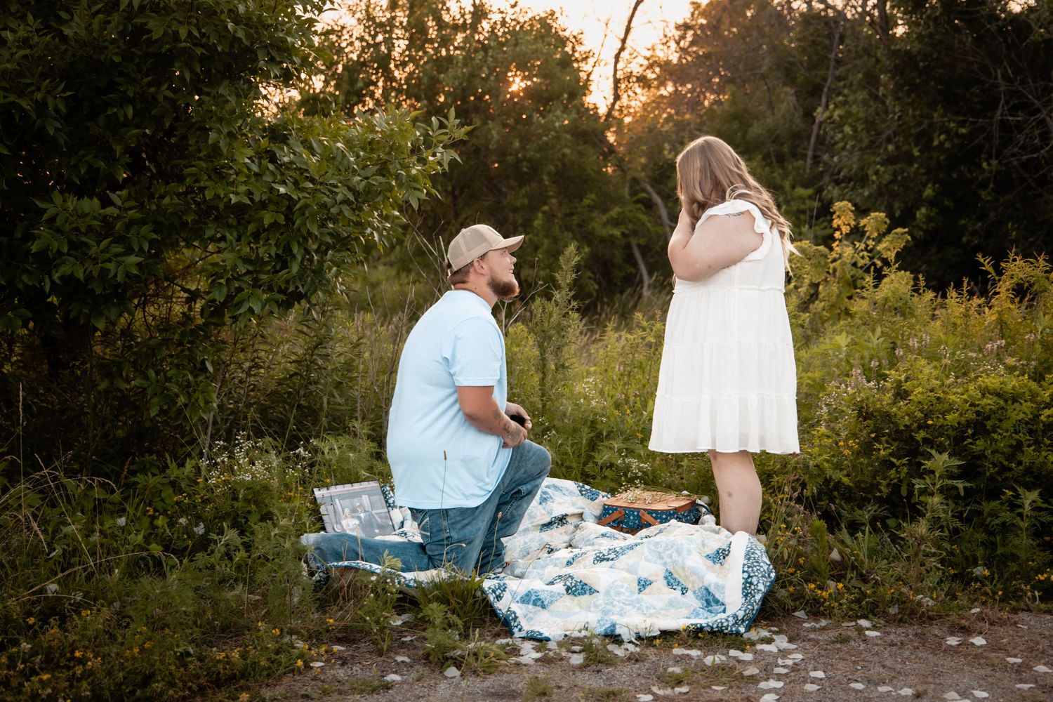 Man proposing on one knee to woman in white dress during golden hour in a lush green outdoor setting.