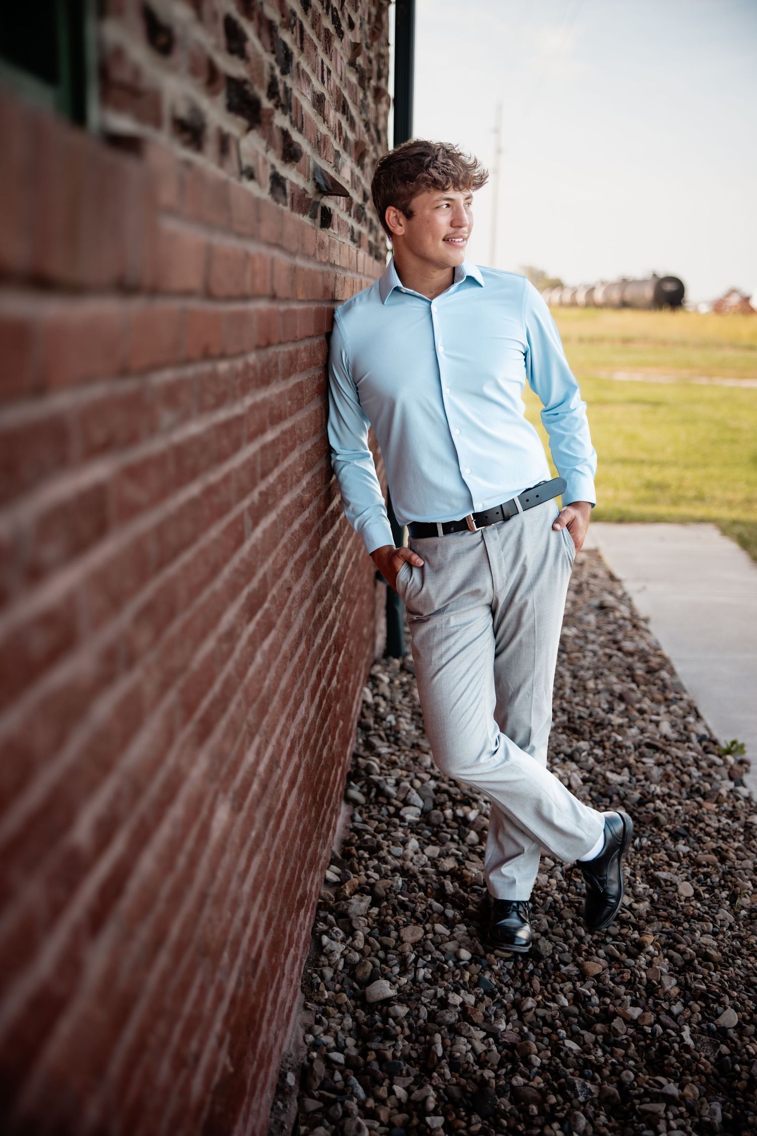 Young man in light blue dress shirt and gray slacks leaning against a red brick wall outdoors.