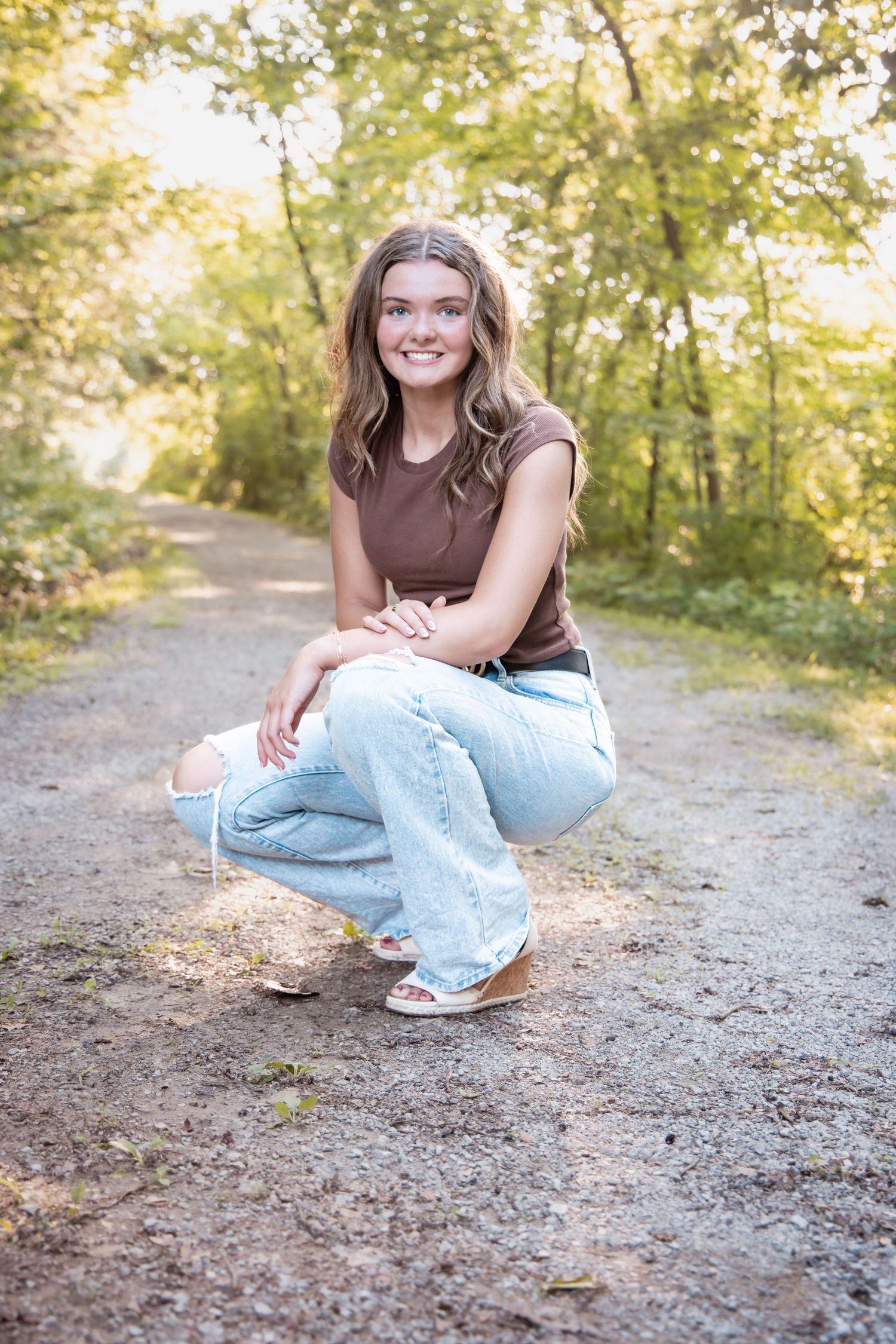 Young woman in brown top and flare jeans crouching on a forest path, smiling outdoors.