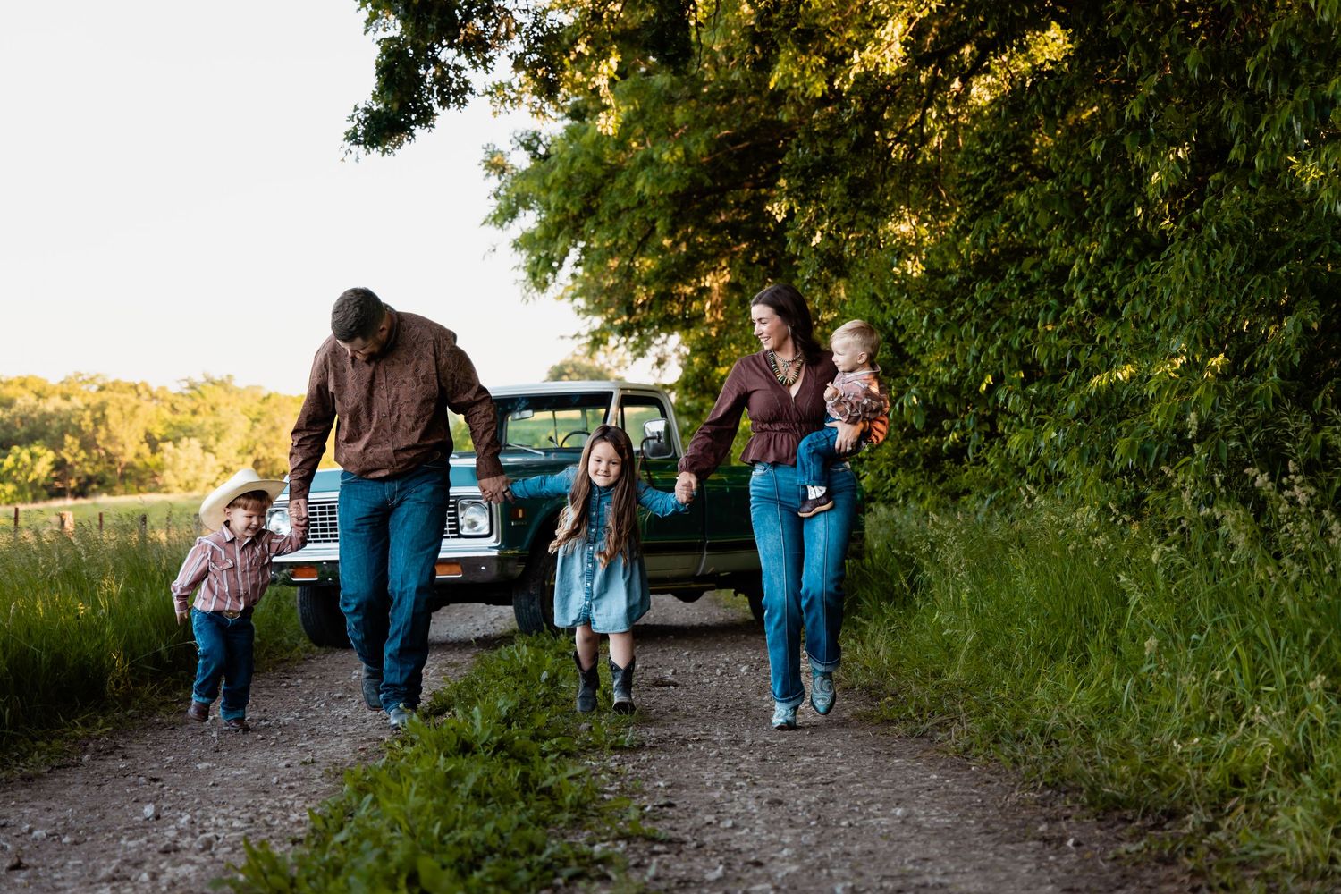 A joyful family of five walks along a rural dirt path beside a classic teal pickup truck.
