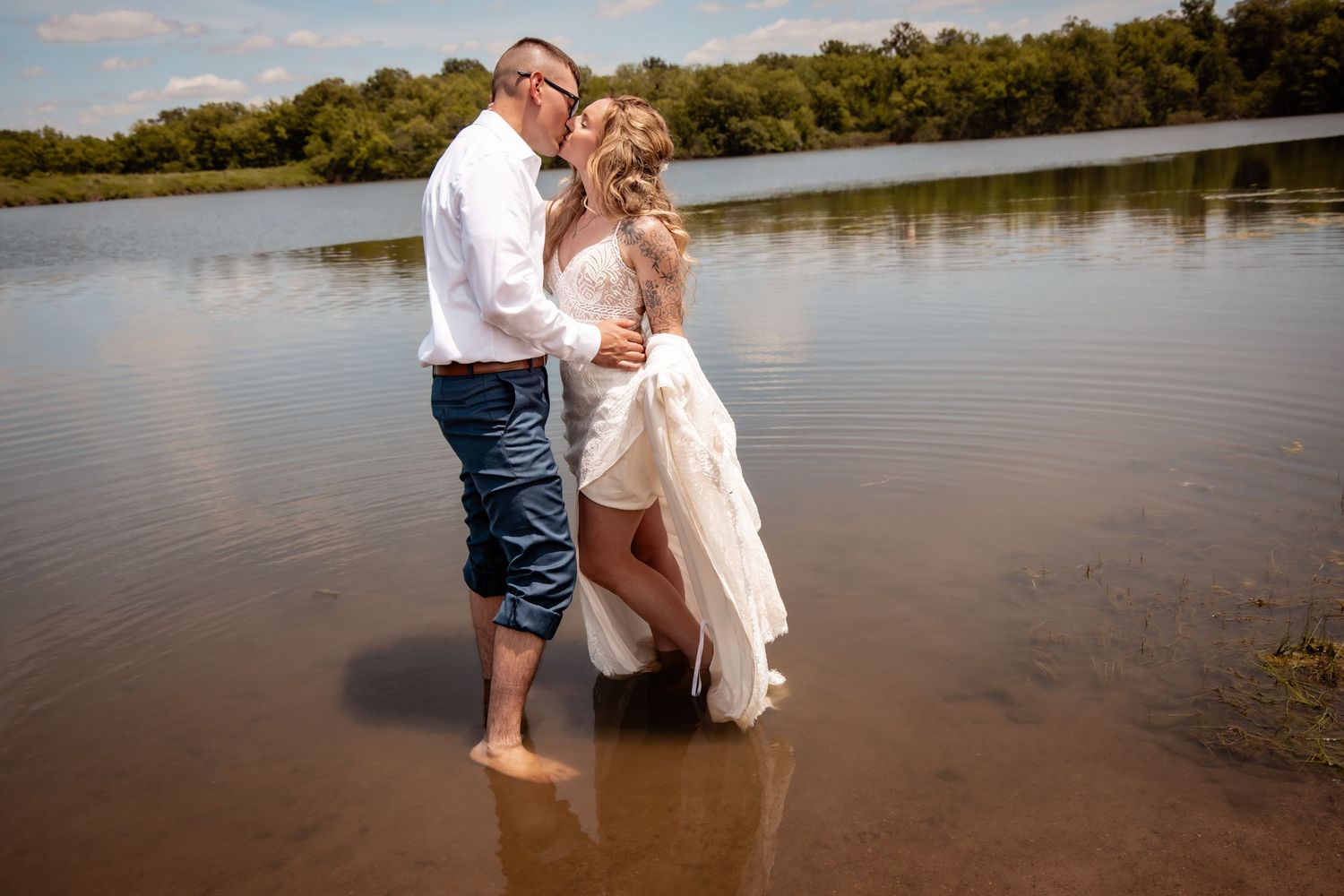 Bride and groom standing barefoot in shallow water, sharing a romantic kiss in their wedding attire outdoors.