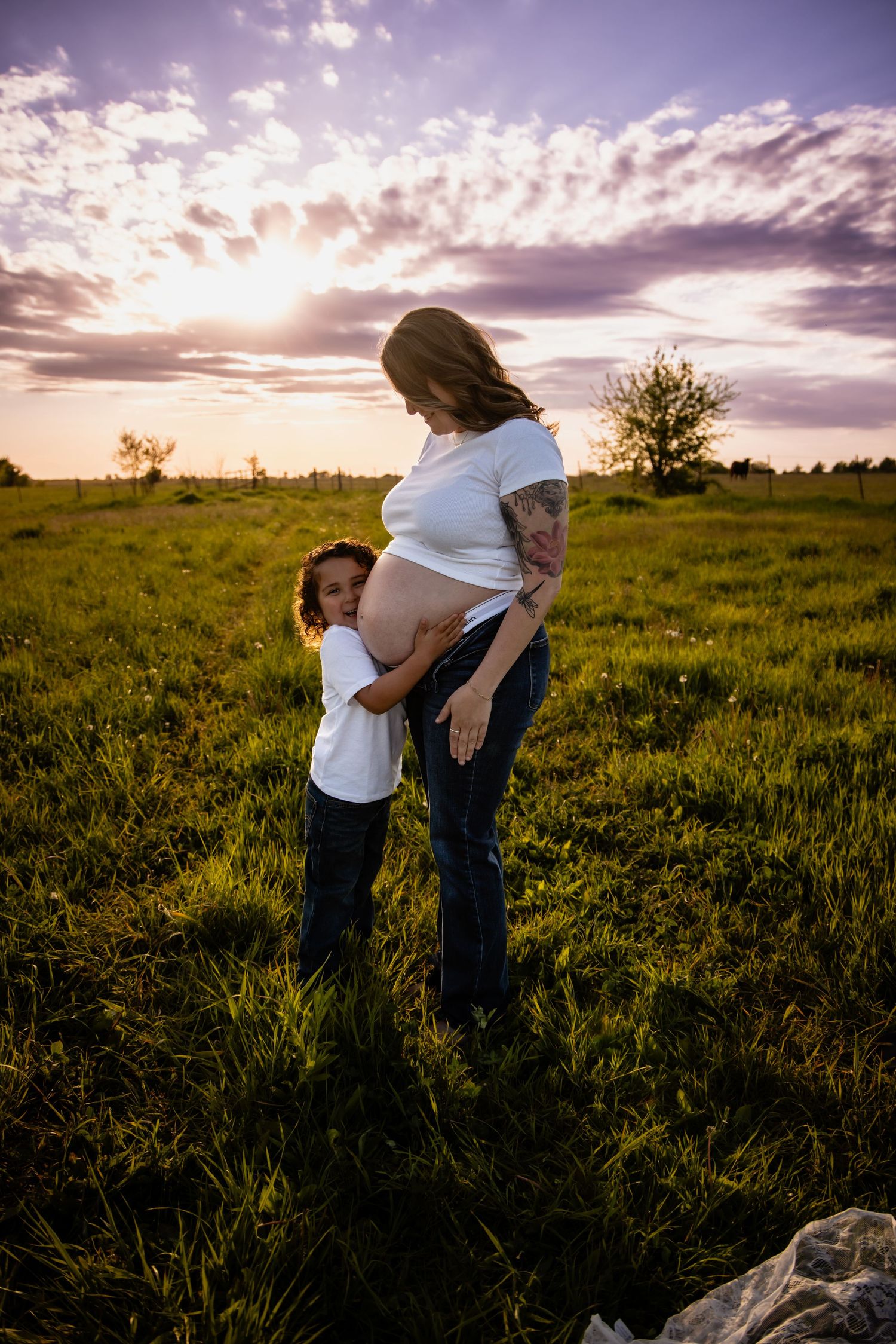 Pregnant woman and child embrace in a green field at golden hour sunset, both wearing white shirts.