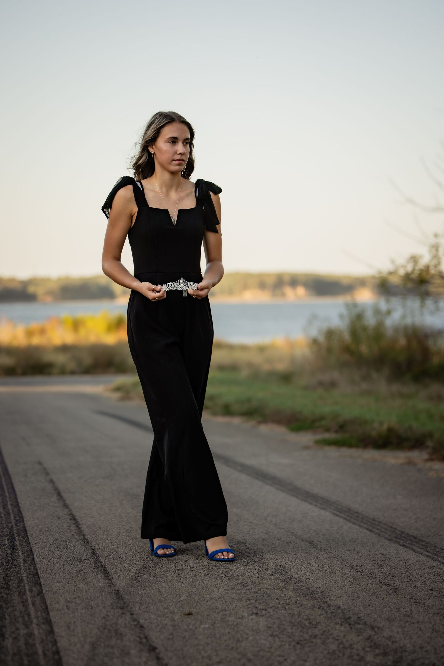 Woman in black sleeveless jumpsuit with bow shoulders and jeweled belt stands on road at sunset.