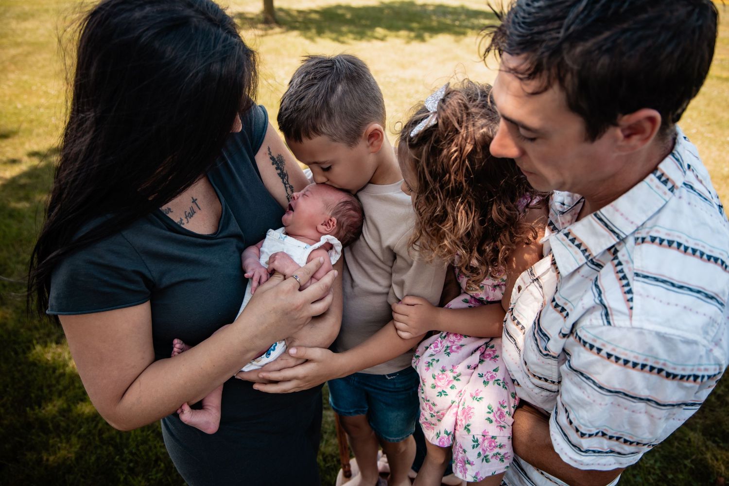 Family of five gathering outdoors, parents holding newborn baby while older siblings lean in to give kisses.